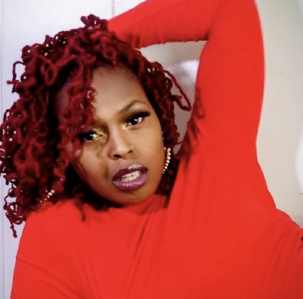 Woman with curly red hair wearing a red top, posing with her arm behind her head, looking at the camera.
