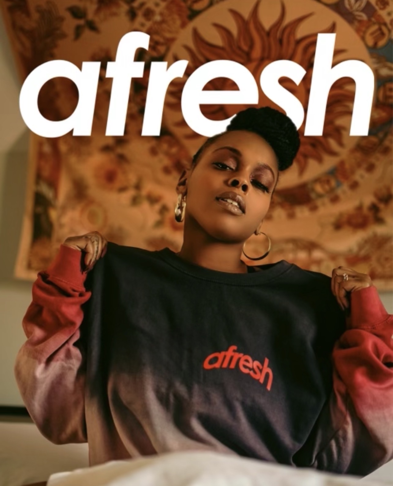 A woman with short hair and earrings wears a black sweatshirt with red text, standing in front of a patterned background.