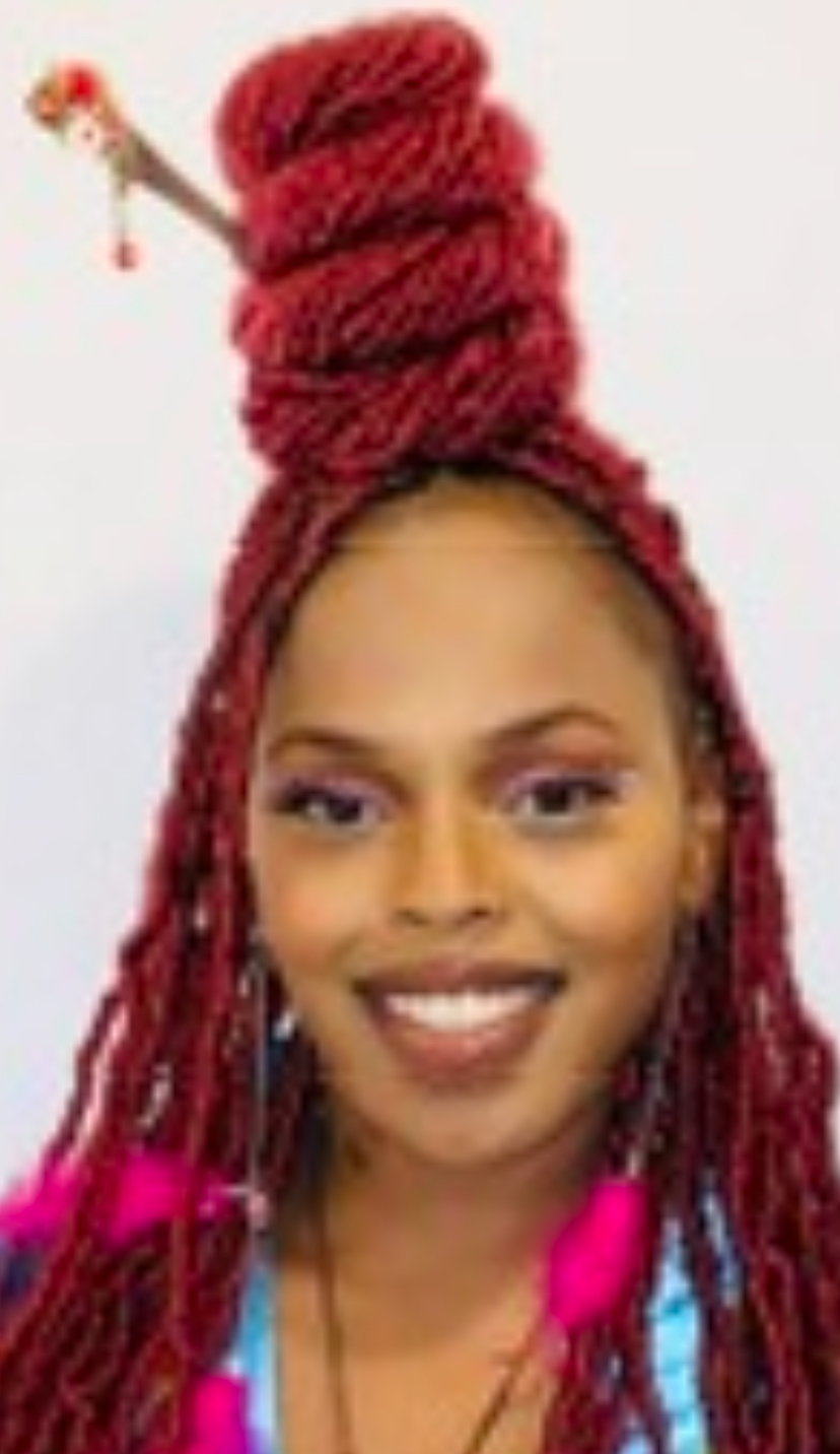Woman with red dreadlocks styled in a tall, coiled bun, smiling, wearing earrings and a blue top.