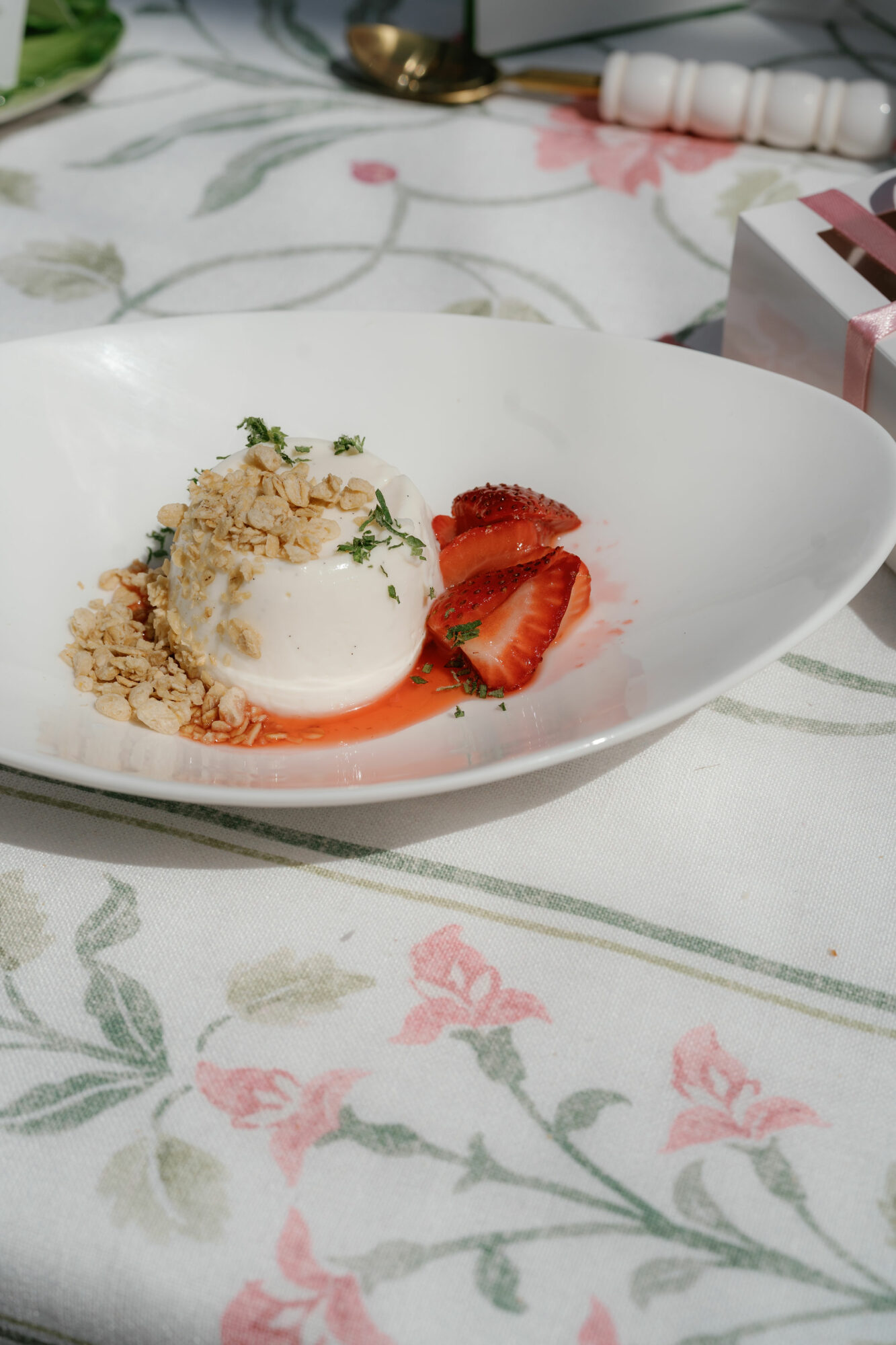 Dessert with sliced strawberries, cream, and crumble on a white plate, on a floral tablecloth.