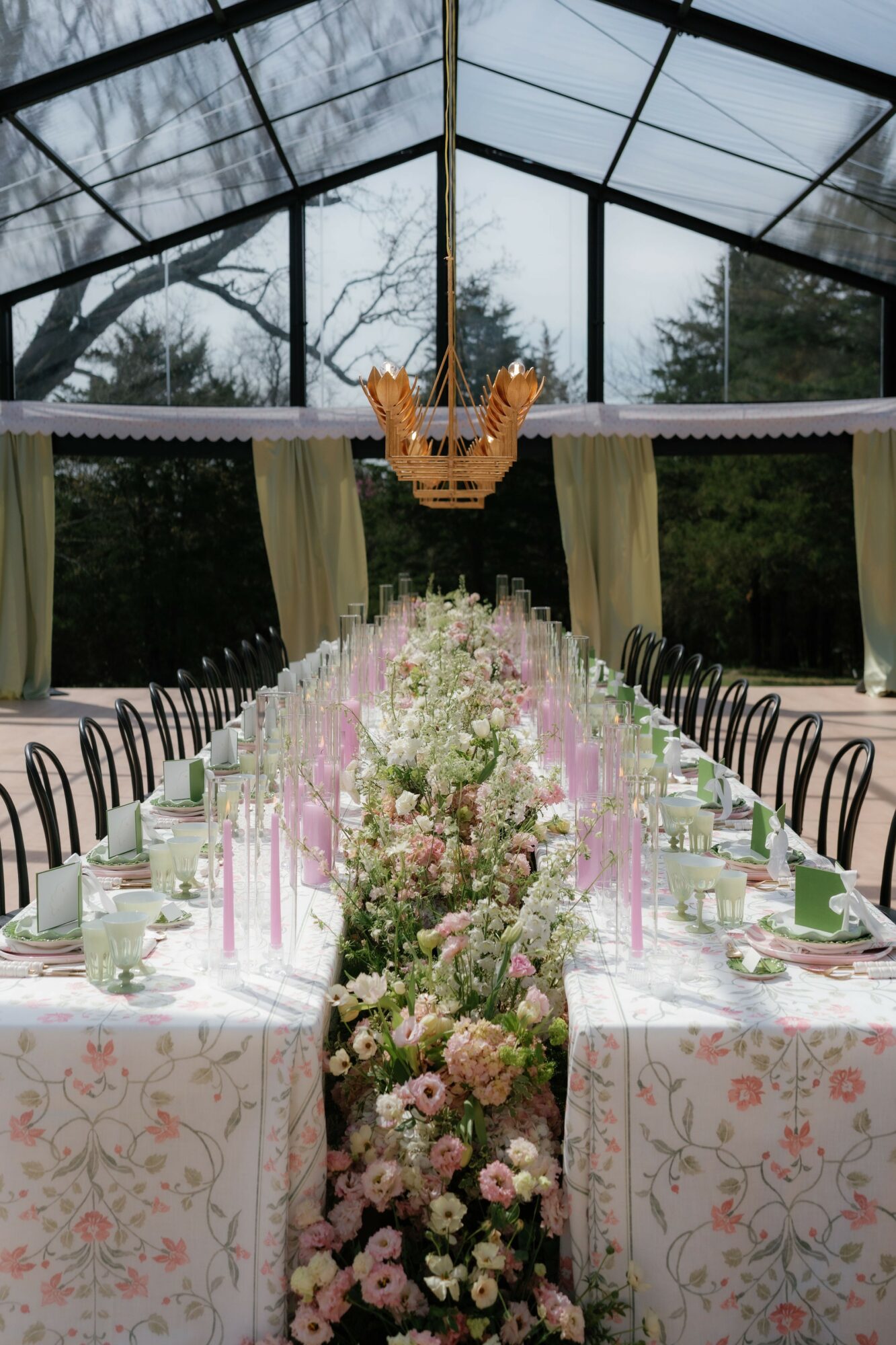 Long dining table decorated with flowers and candles inside a glass conservatory, with a crown-shaped decoration hanging above.