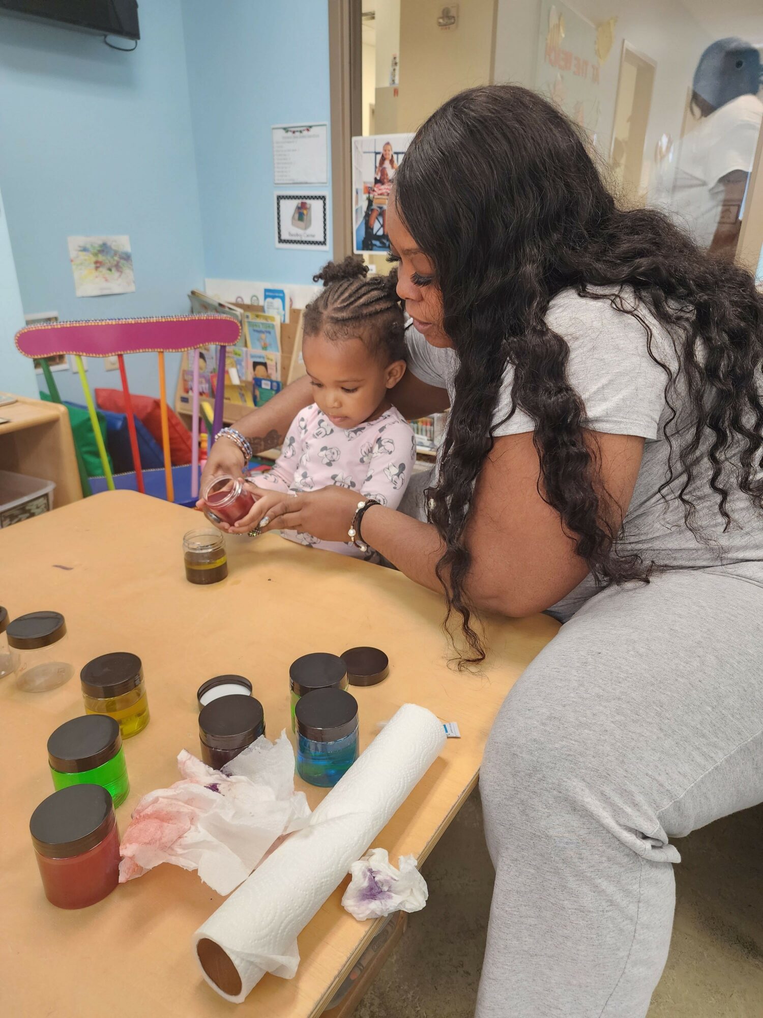 Two children and an adult sitting at a table with art supplies, including paint jars and paper rolls.