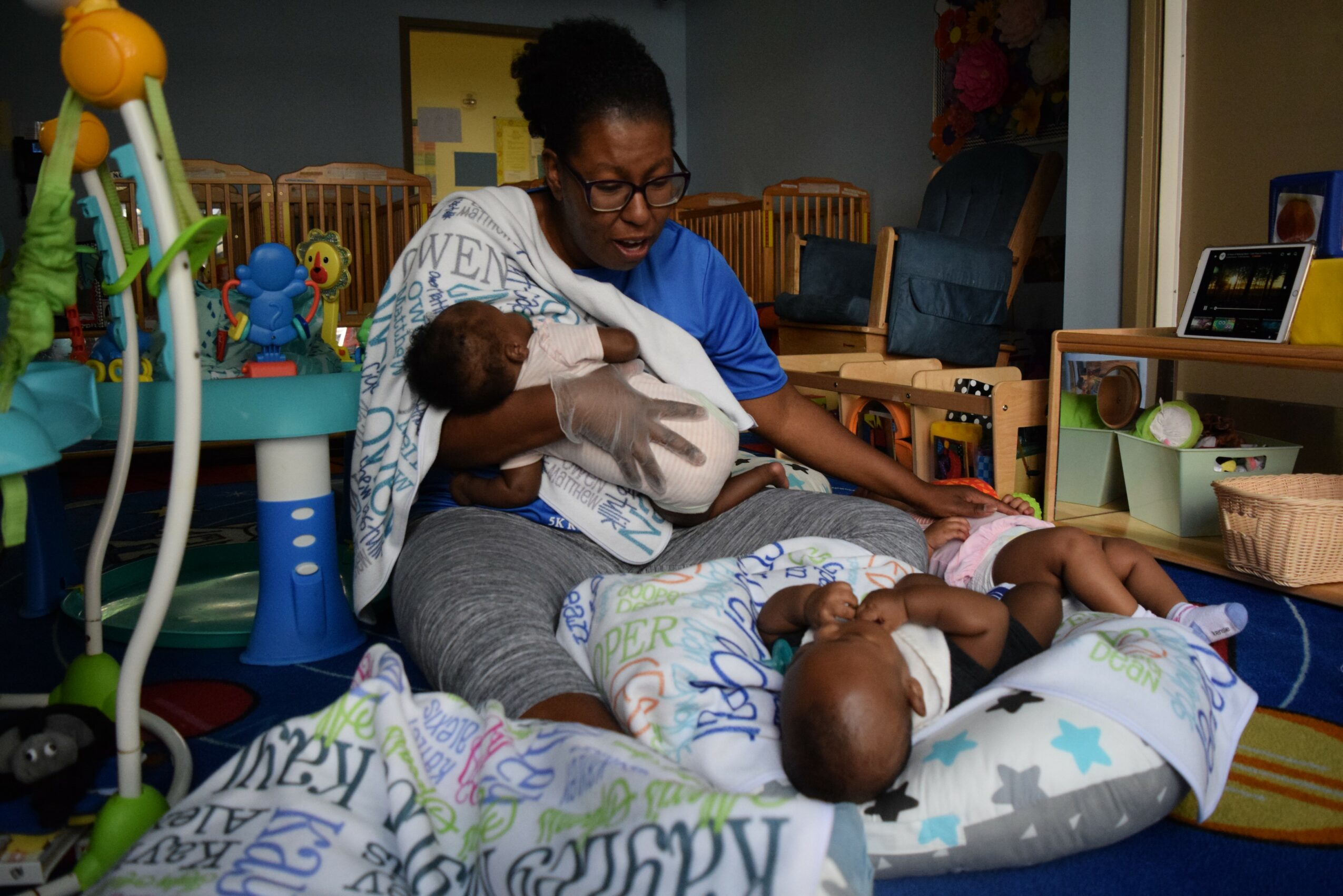 Woman breastfeeding two babies in a playroom with toys and furniture, one baby lying on a pillow, the other in her arms.