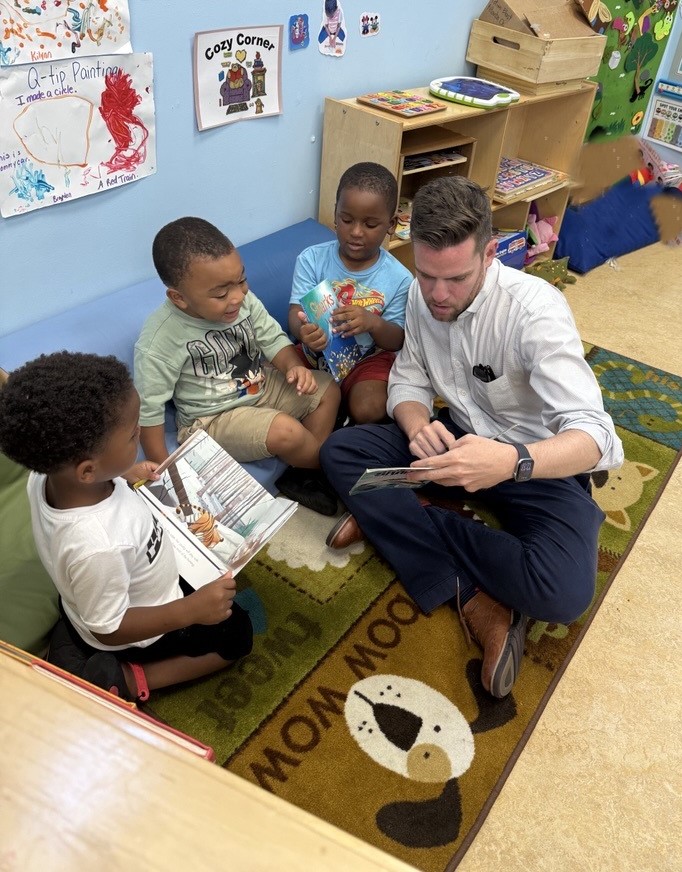 Adult man sitting on the floor with three children, reading a book together in a classroom, colorful wall decorations, and educational materials.
