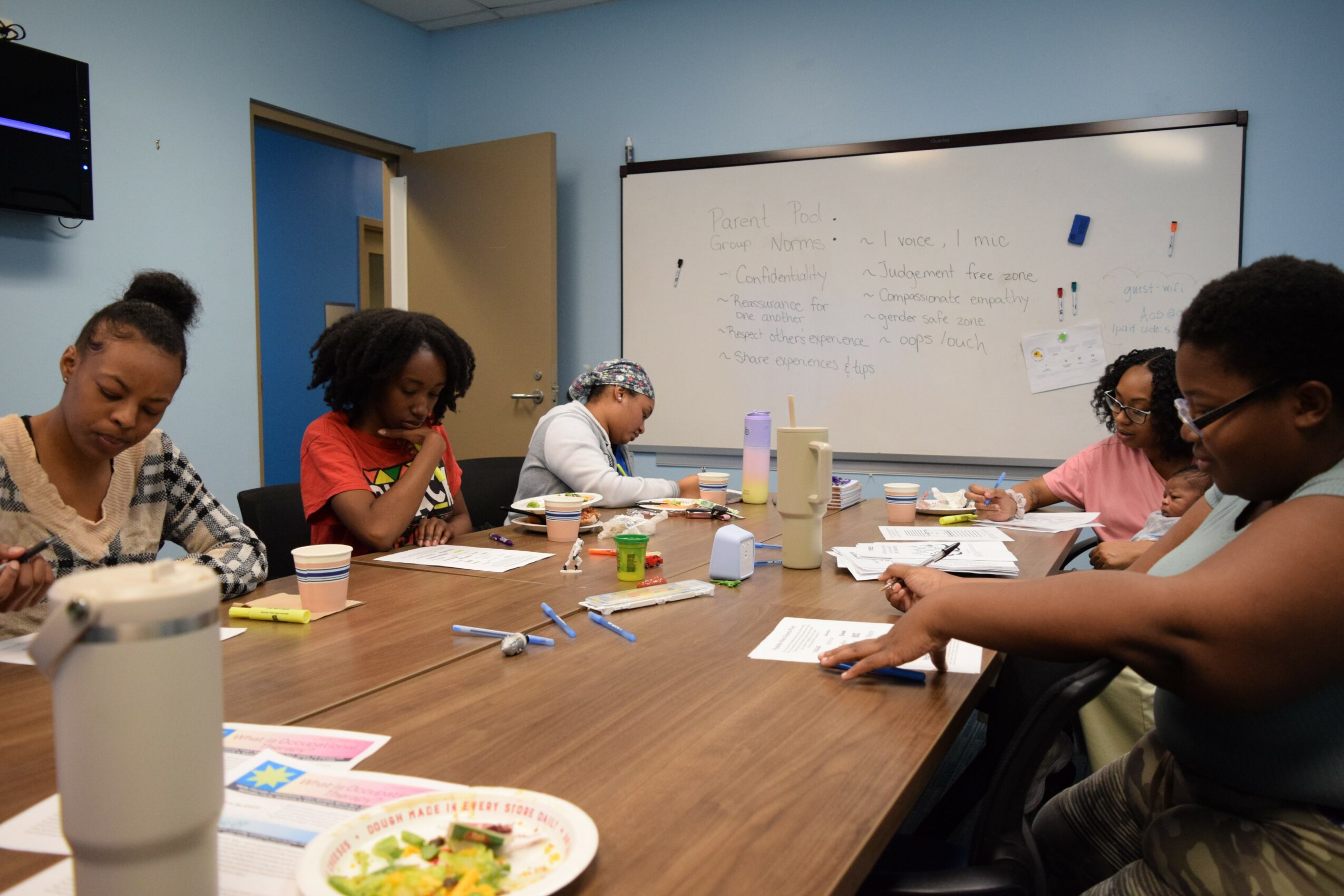Six people sit around a table in a classroom, writing and working on papers, with a whiteboard behind them.