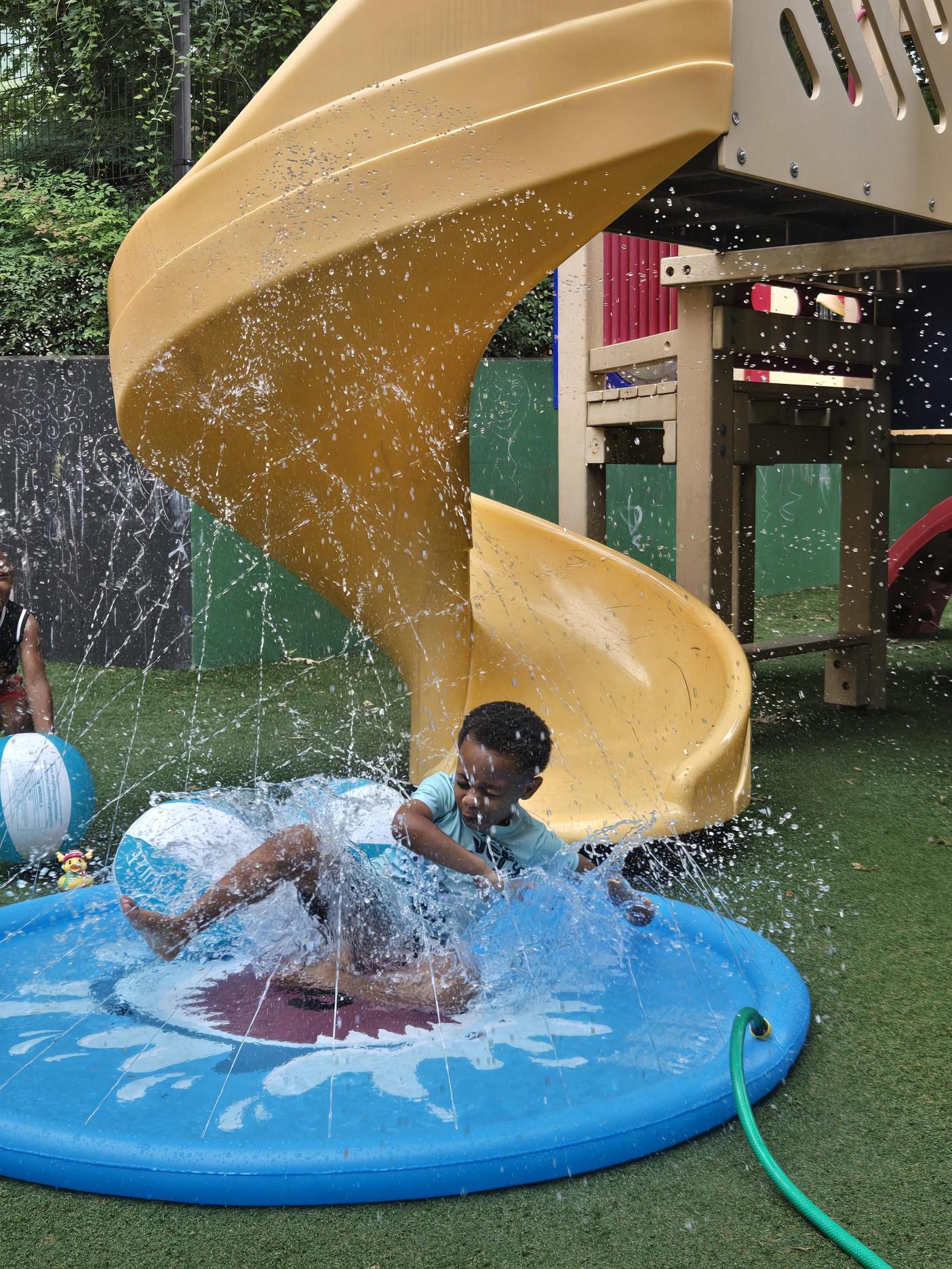 Child playing in water near a yellow slide and blue pool, with water splashing around.