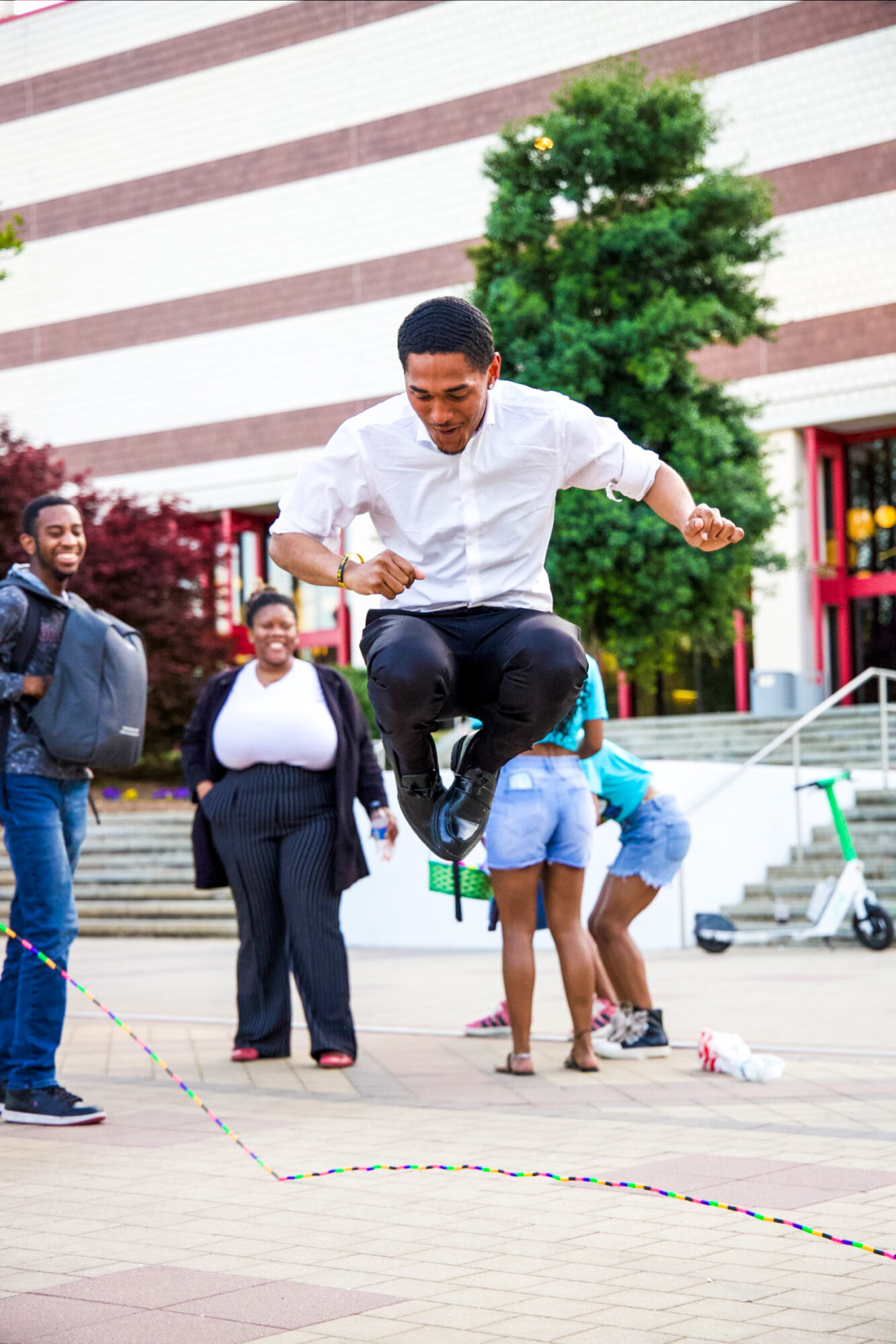 Person jumping over a rope outdoors with onlookers, trees, and a building in the background.