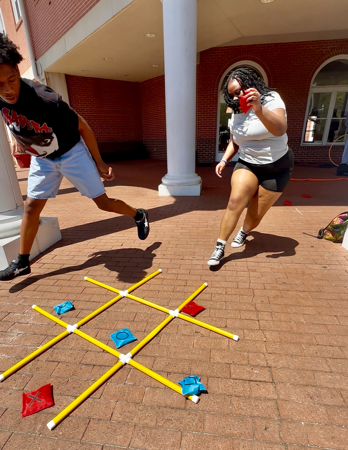 Two people playing a game on brick pavement, with a yellow grid and colored pieces, outside a building with arches.