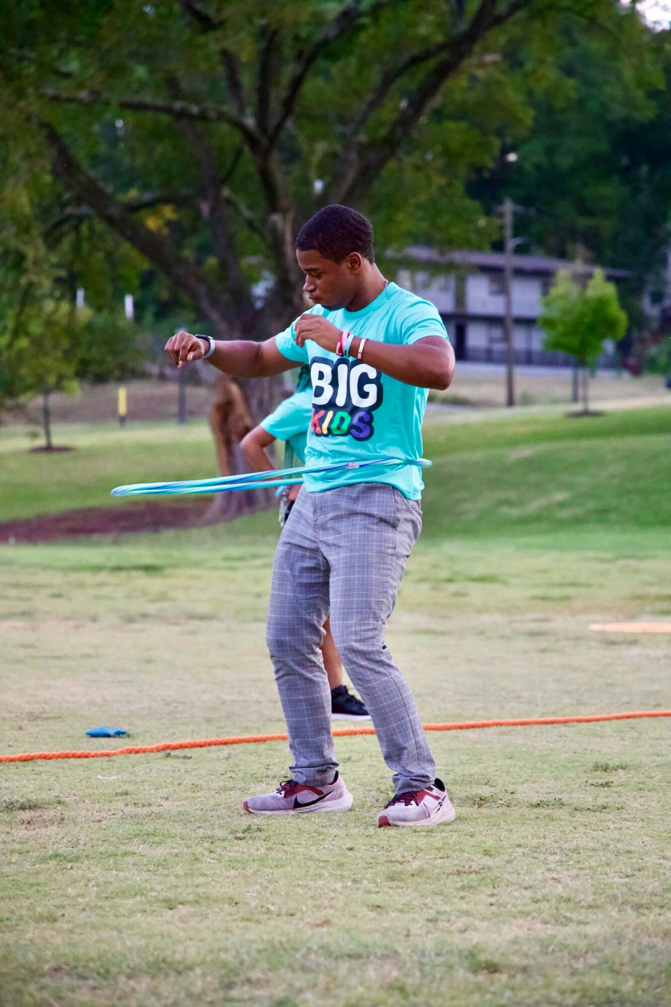 Child playing with a hula hoop outdoors in a park with trees and grass, wearing a blue shirt and gray pants.