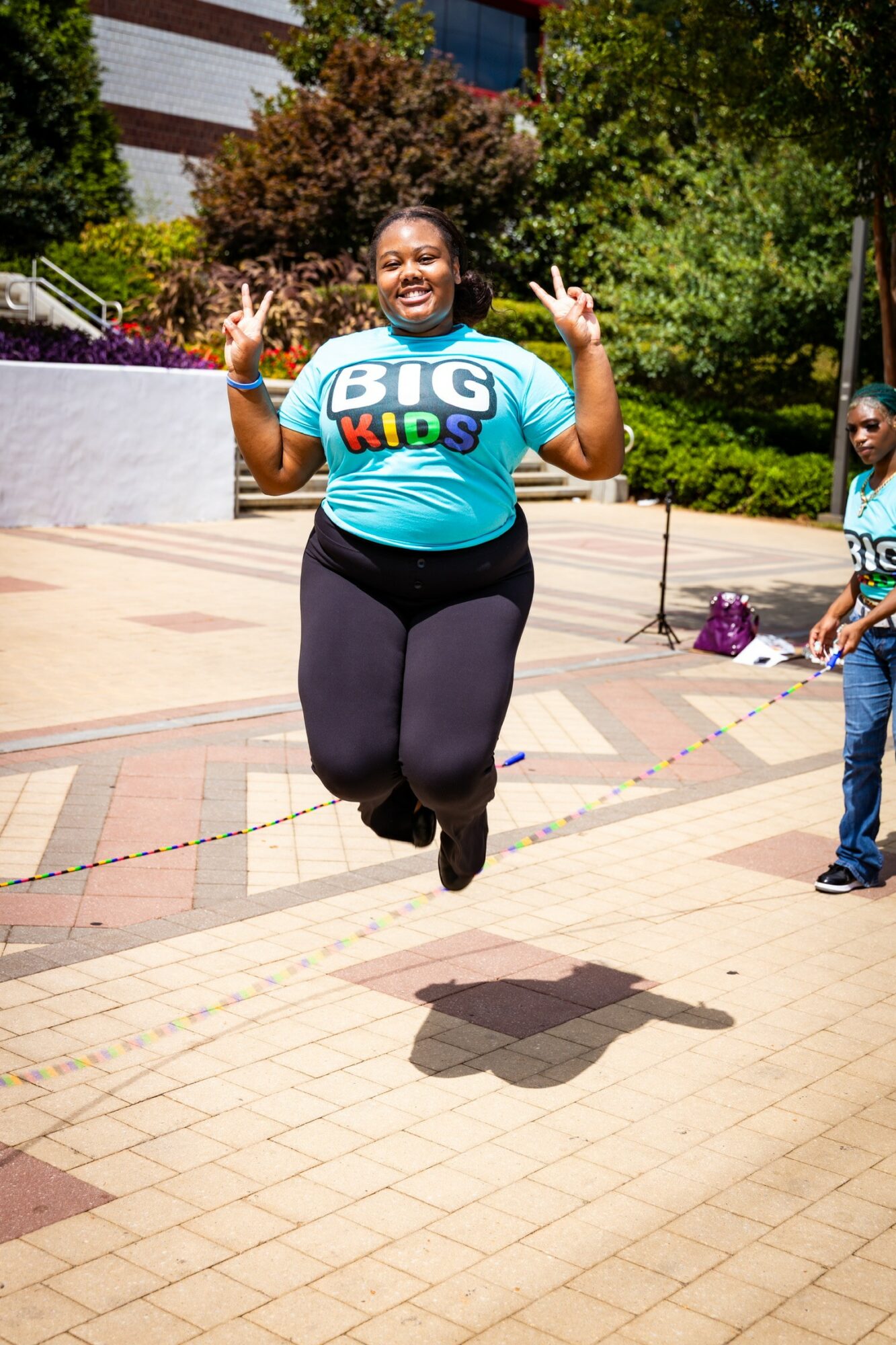 Person jumping with arms raised and peace signs, wearing a blue shirt with 'BIG KIDS' text, outdoors on a sunny day.