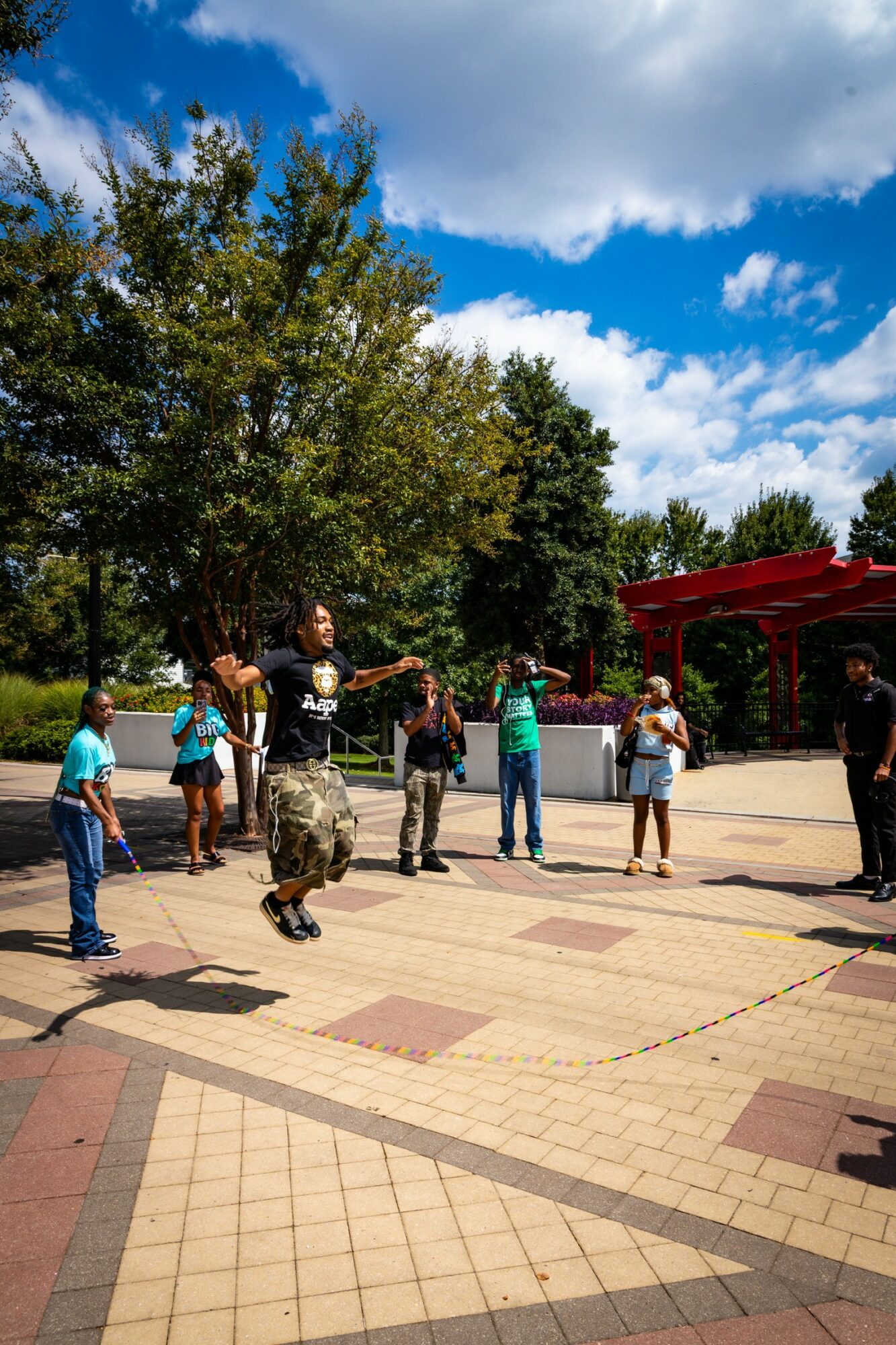 Person jumping with arms raised outdoors, surrounded by children and adults, trees and a red structure in the background.