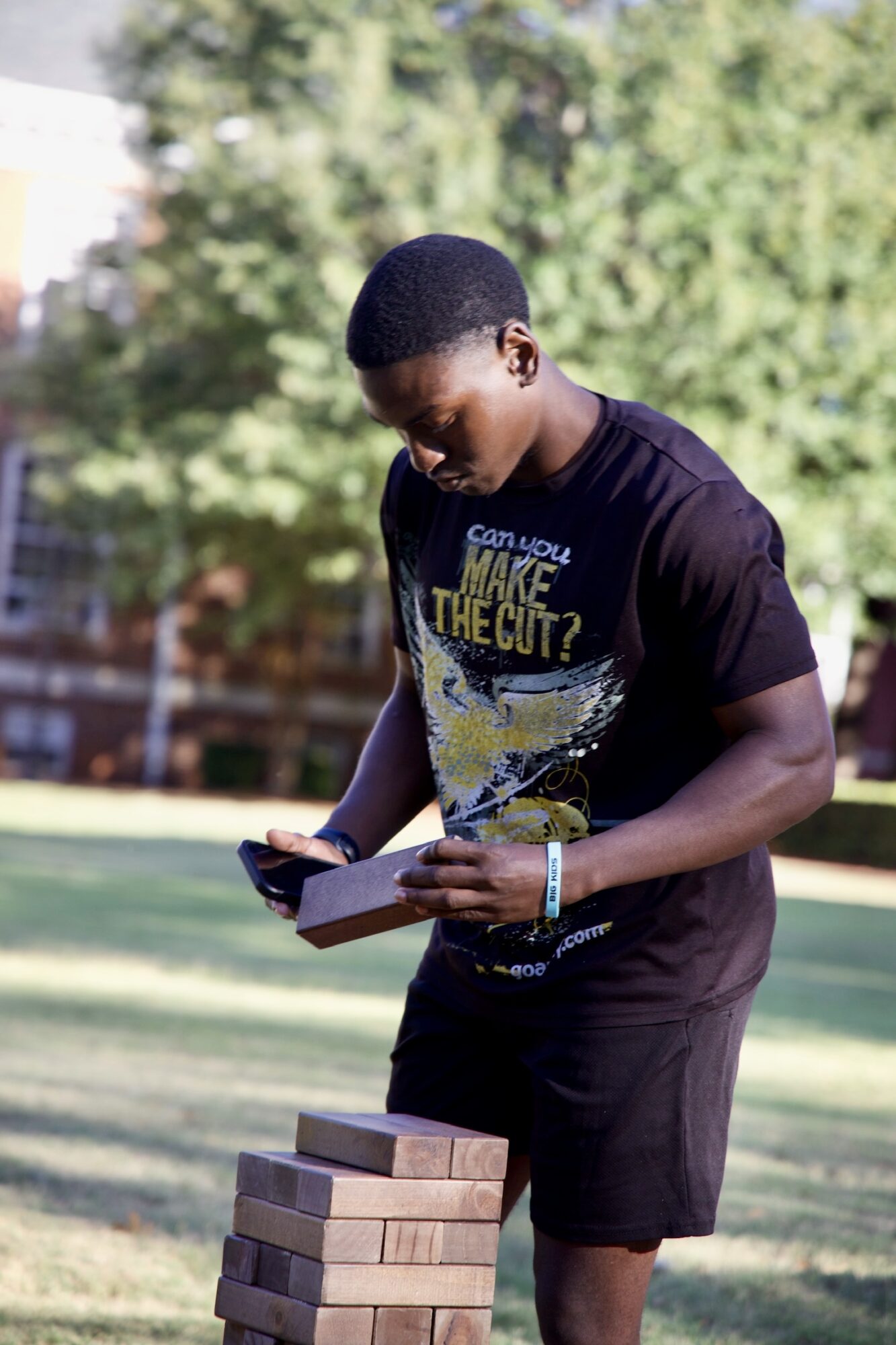 Young man outdoors, reading a book, with a stack of bricks nearby, trees and a fence in the background.