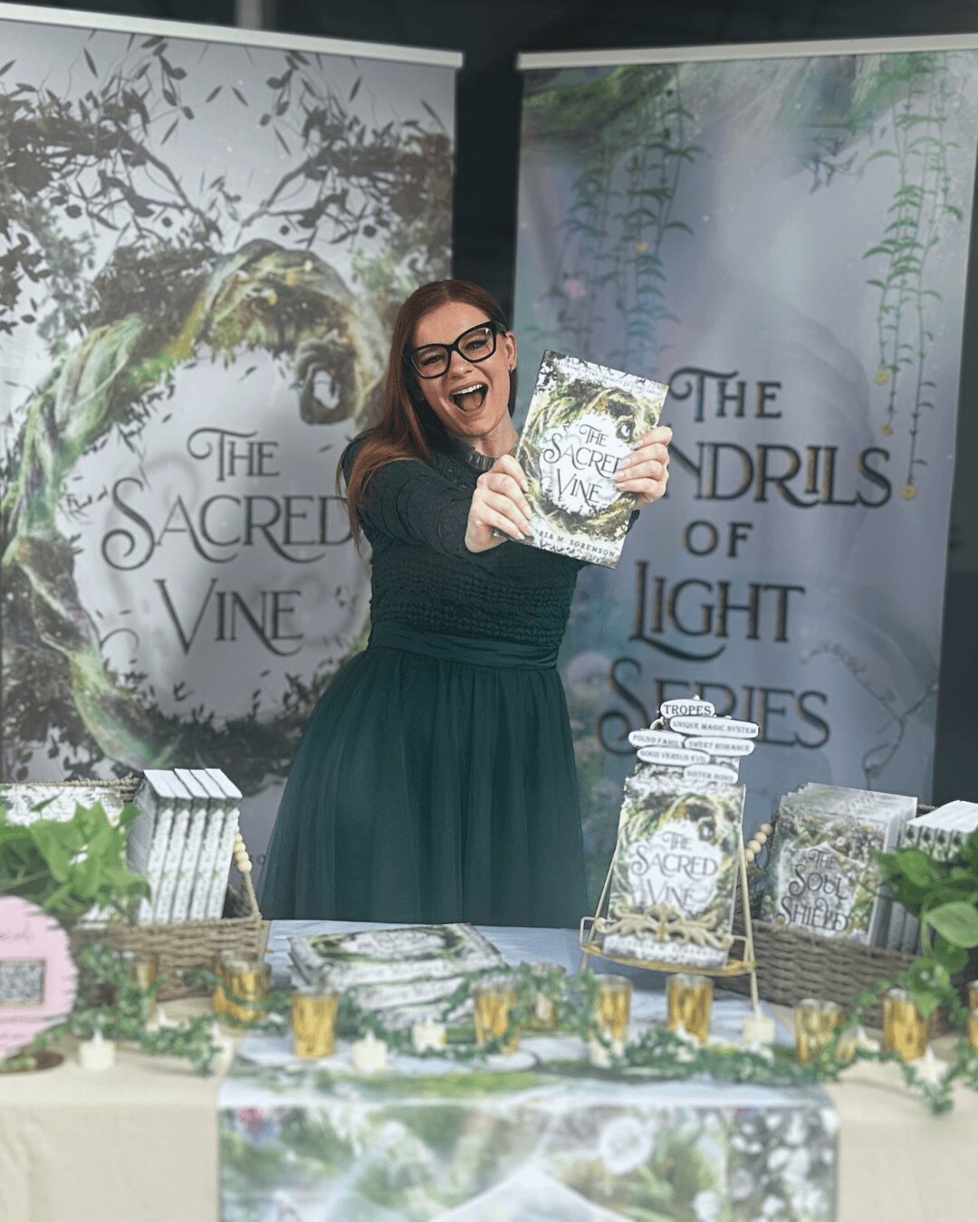Woman smiling and holding a book at a table with books and decorations, two banners in the background, one with a tree and the other with hanging plants.