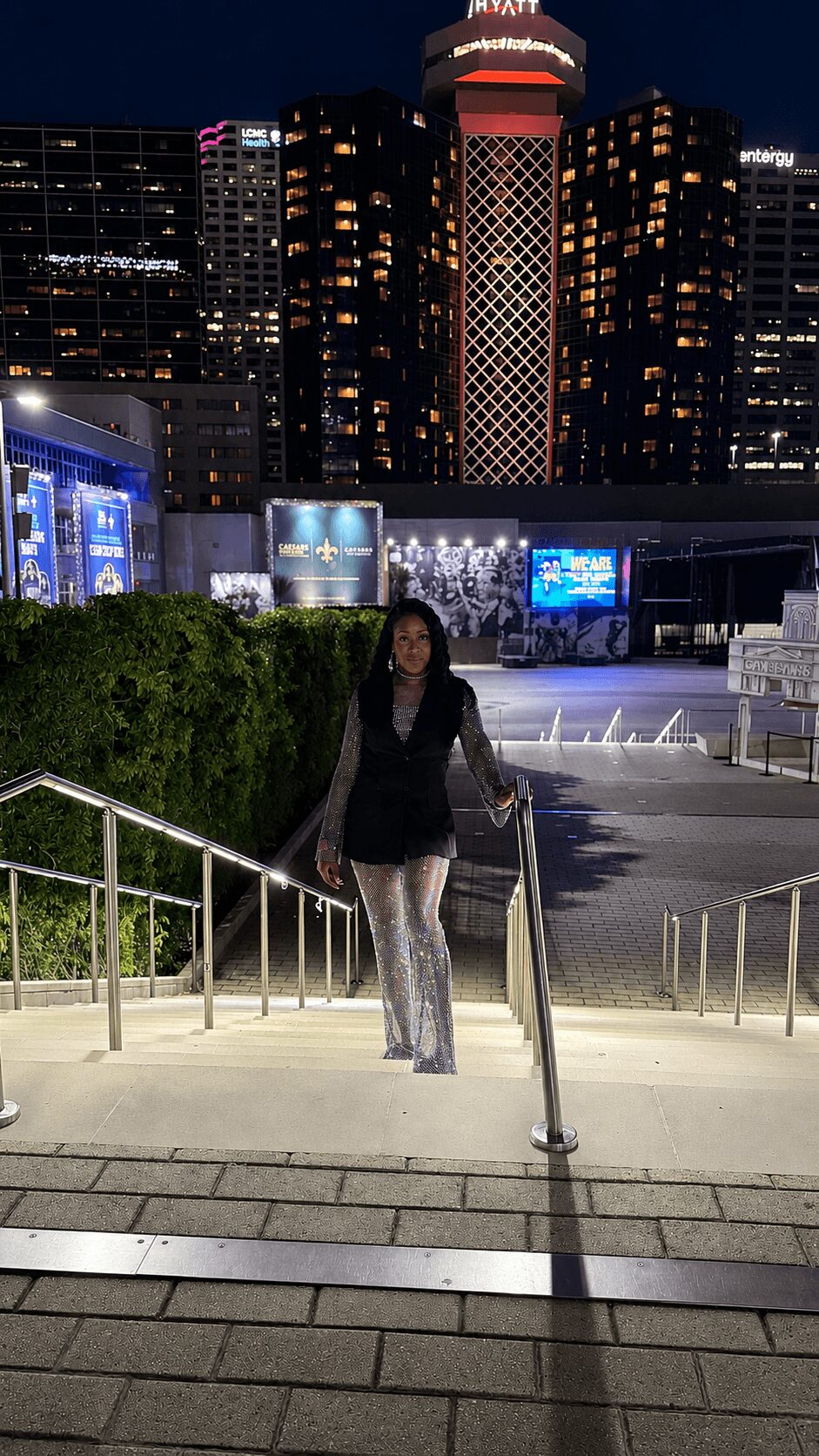 Person standing on stairs at night with city skyline and illuminated tower behind, greenery on side, railing in foreground.