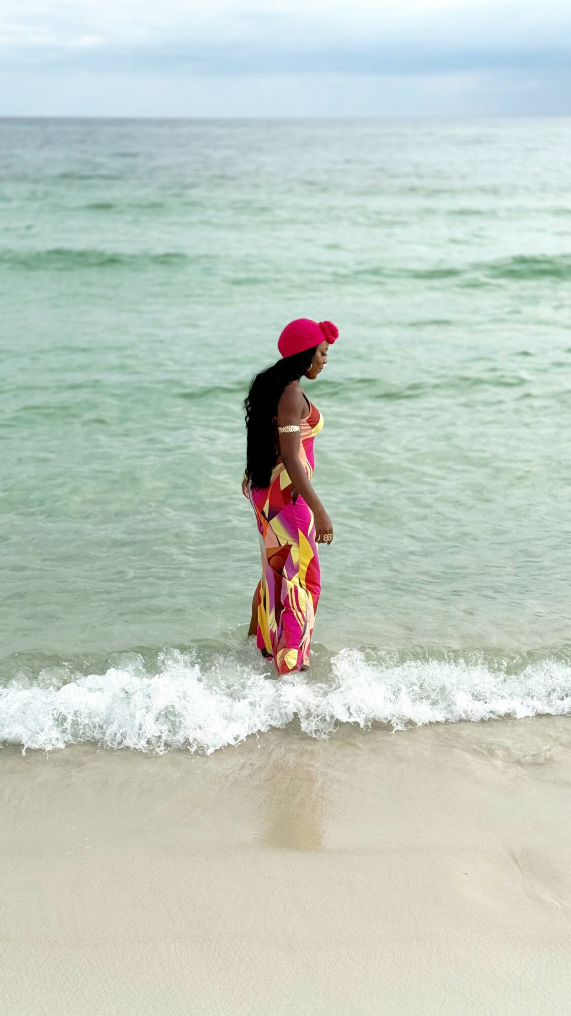 Woman in pink hat and colorful dress standing in ocean waves at the beach.