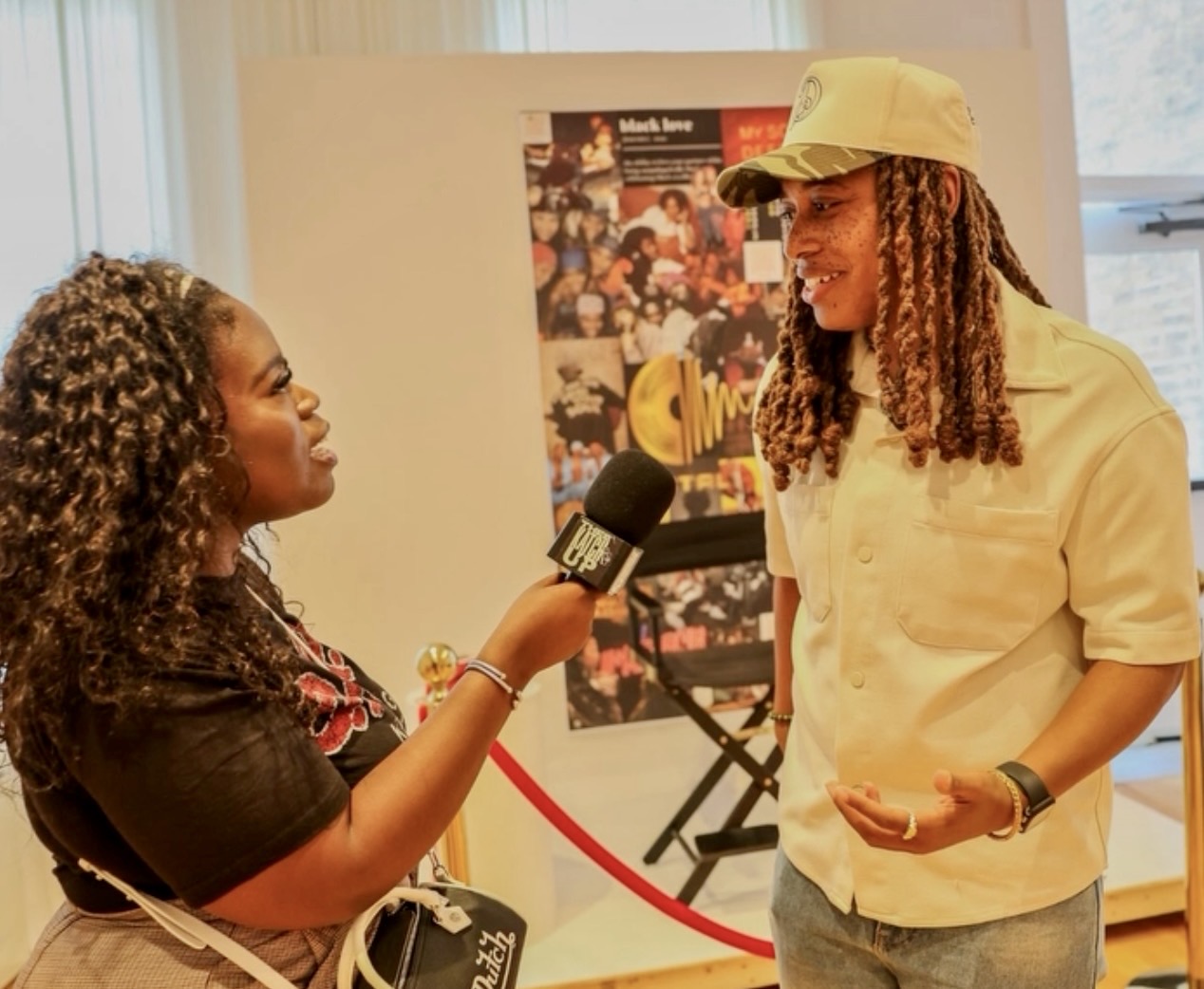 Two women engaged in an interview, one holding a microphone, in an indoor setting with a poster in the background.