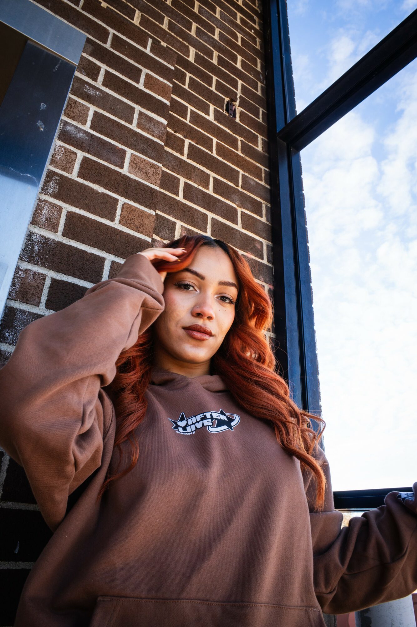 Young woman with long, wavy hair and a brown sweatshirt standing near a brick wall and window, looking at the camera.
