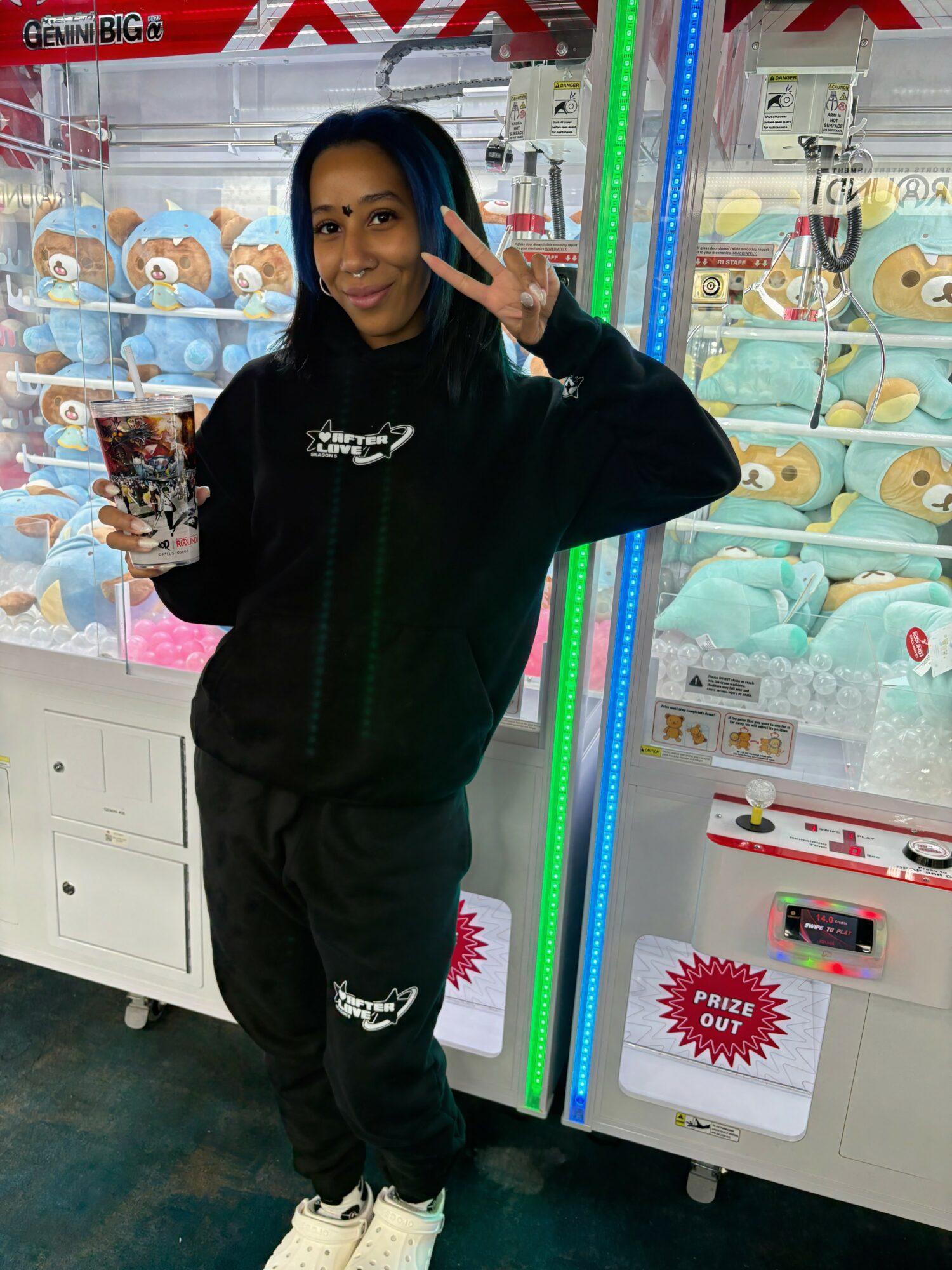 Young woman with dark hair smiling and making a peace sign in front of claw machines filled with plush toys.