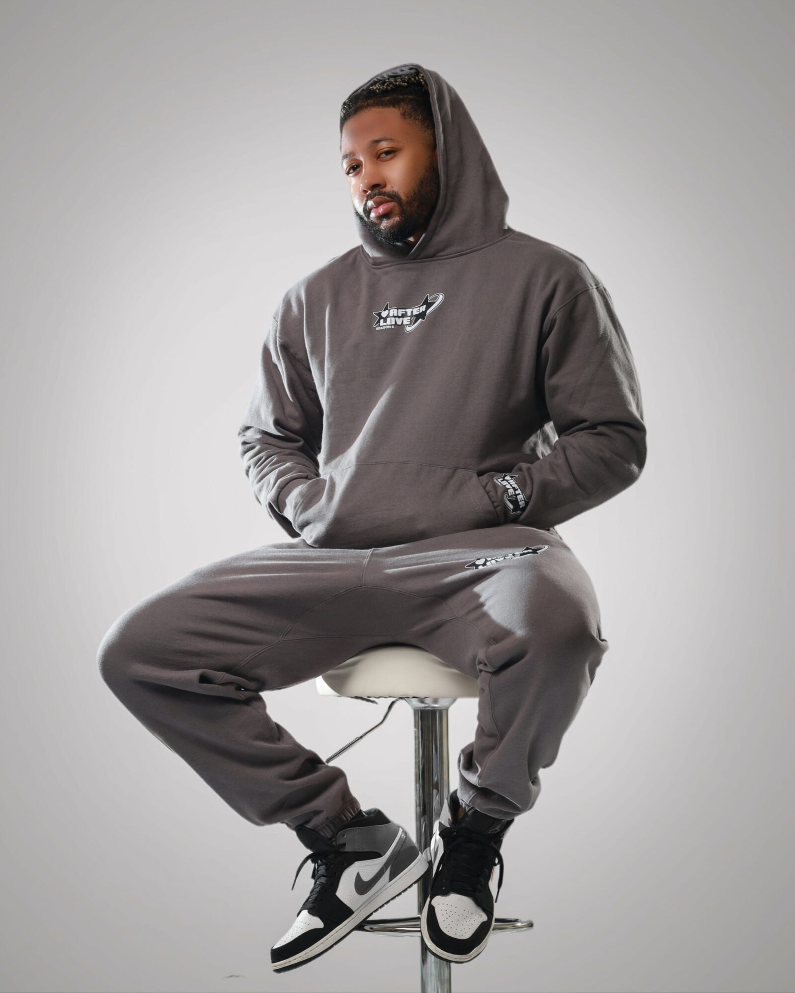 Man with a beard wearing a dark hoodie and sweatpants, sitting on a stool against a plain background.