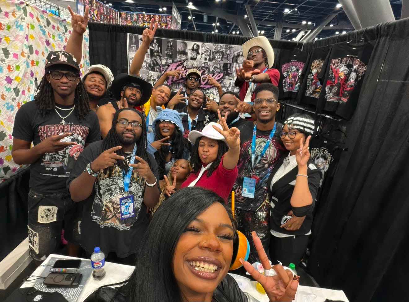 Group of people smiling and posing at an indoor event, some making peace signs, with colorful displays in the background.