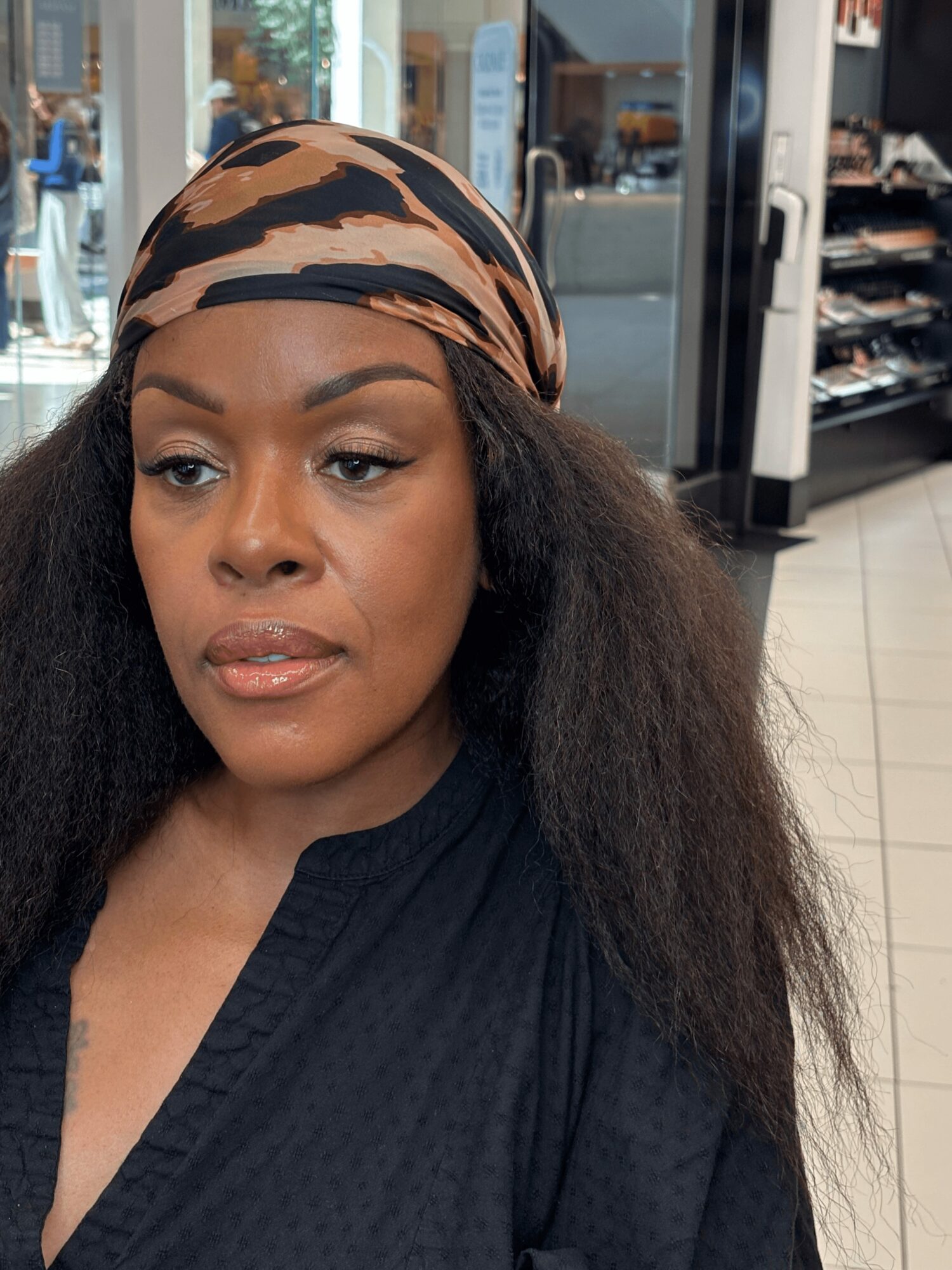 Woman with a patterned headscarf and long hair in a store, looking to the side, wearing a dark top.