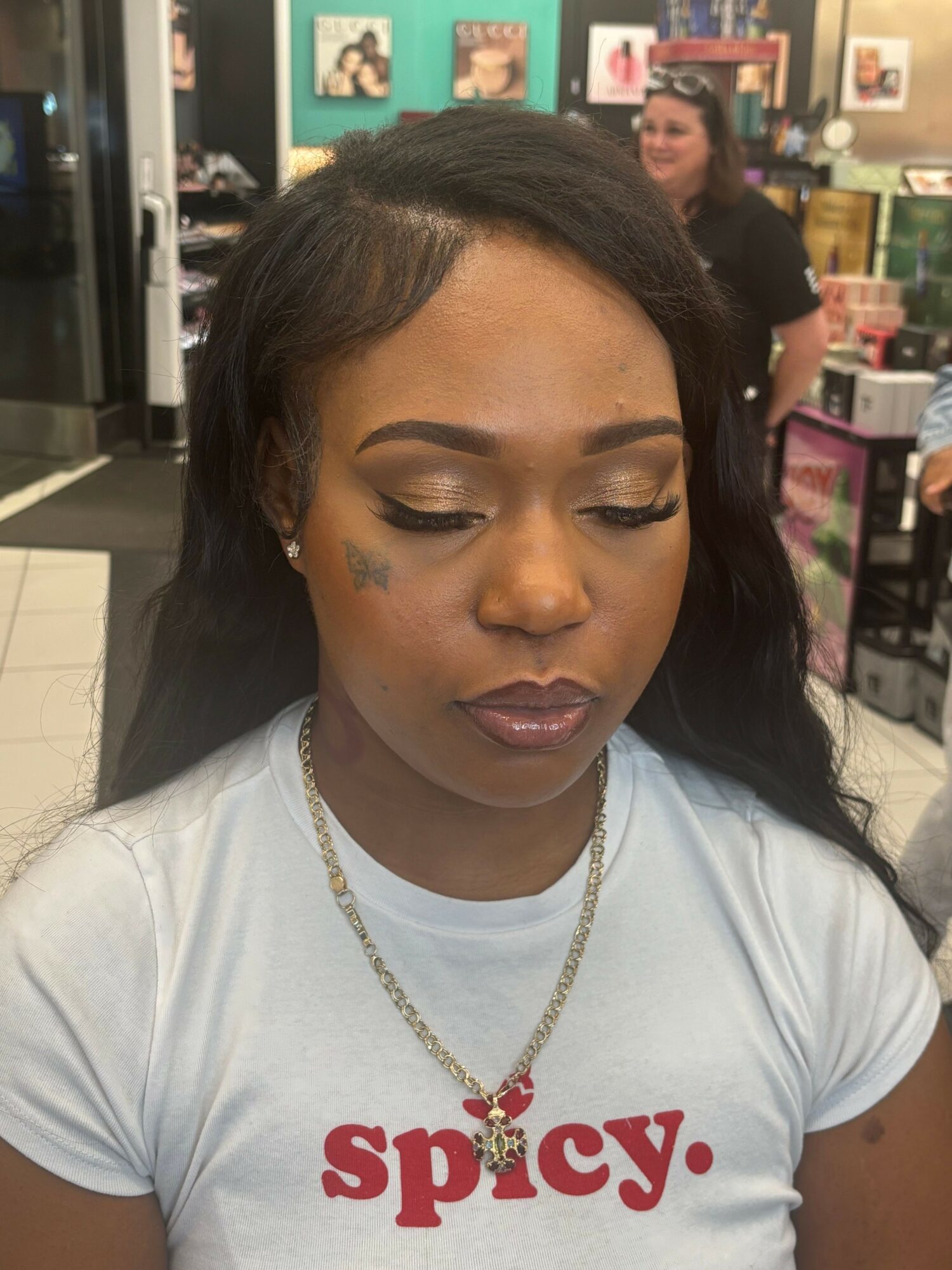 Woman with long dark hair, makeup, and a tattoo on her face, wearing a white t-shirt with red text, in a store.