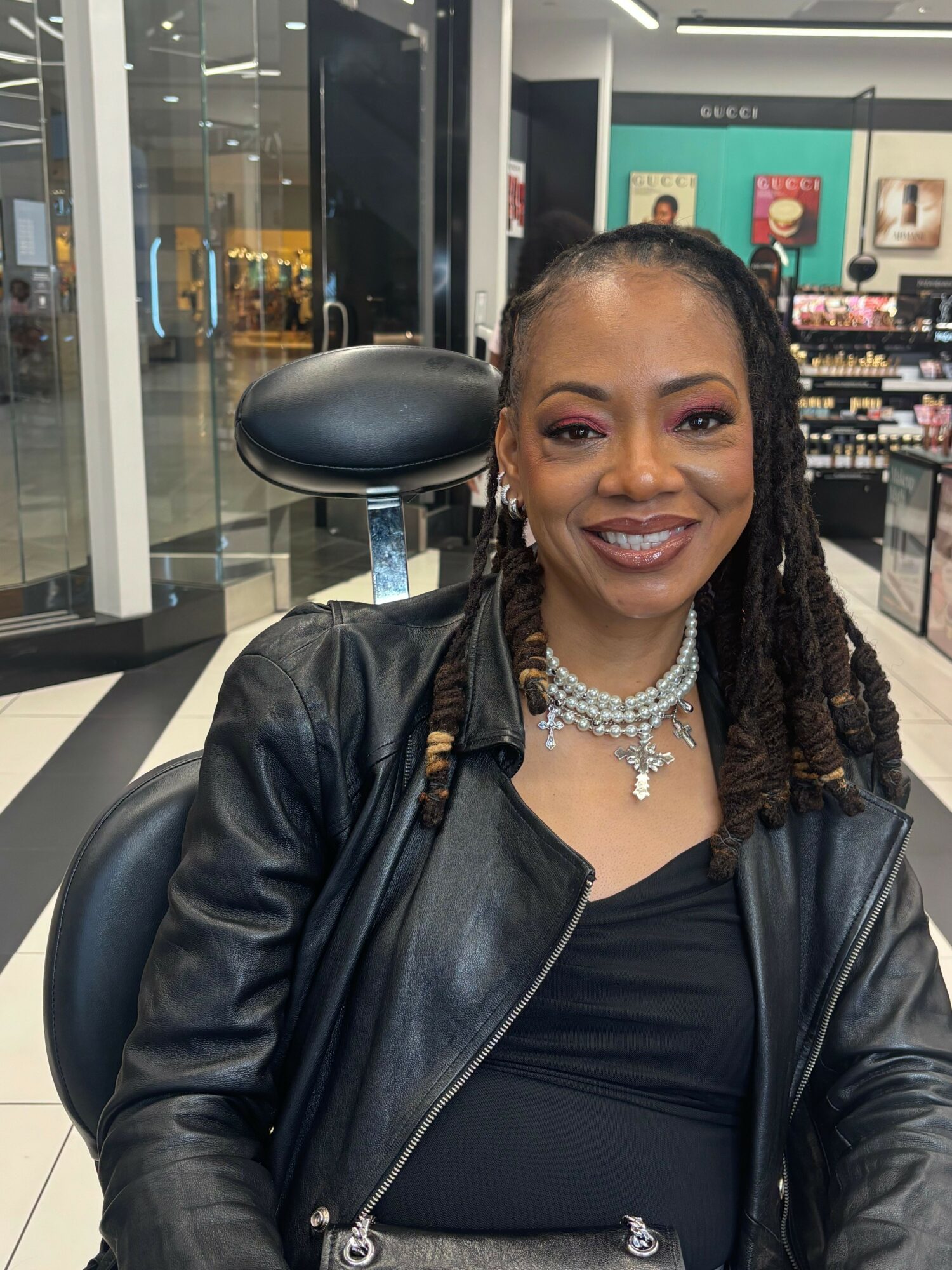 Woman with braided hair, wearing a black leather jacket and pearl necklace, smiling in a store.
