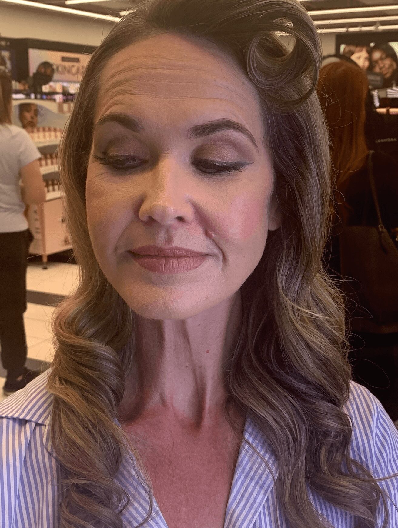 Woman with styled hair and makeup, eyes closed, in a retail store, wearing a striped shirt.