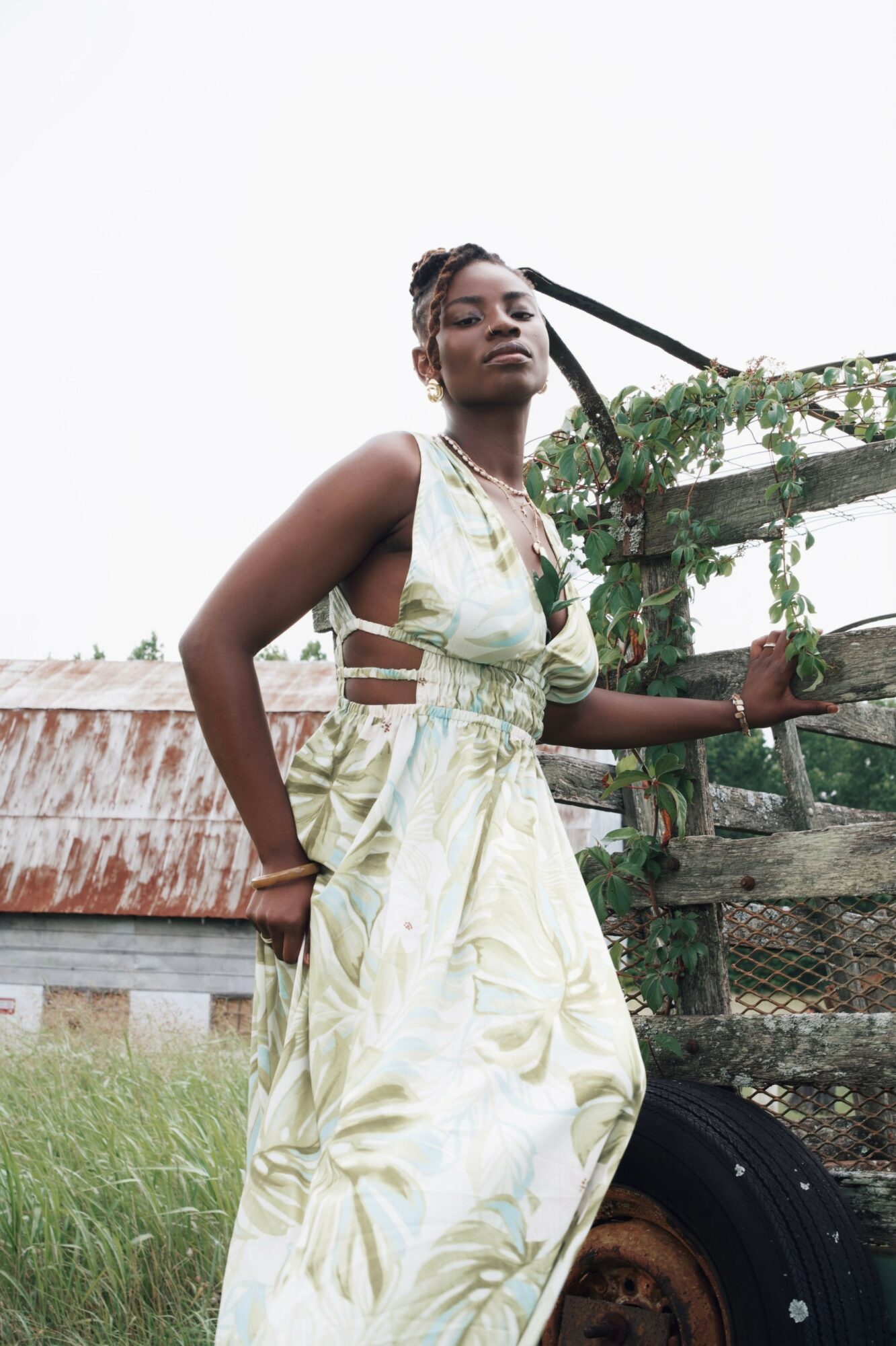 Young woman in a light-colored dress standing outdoors near a wooden fence and greenery.