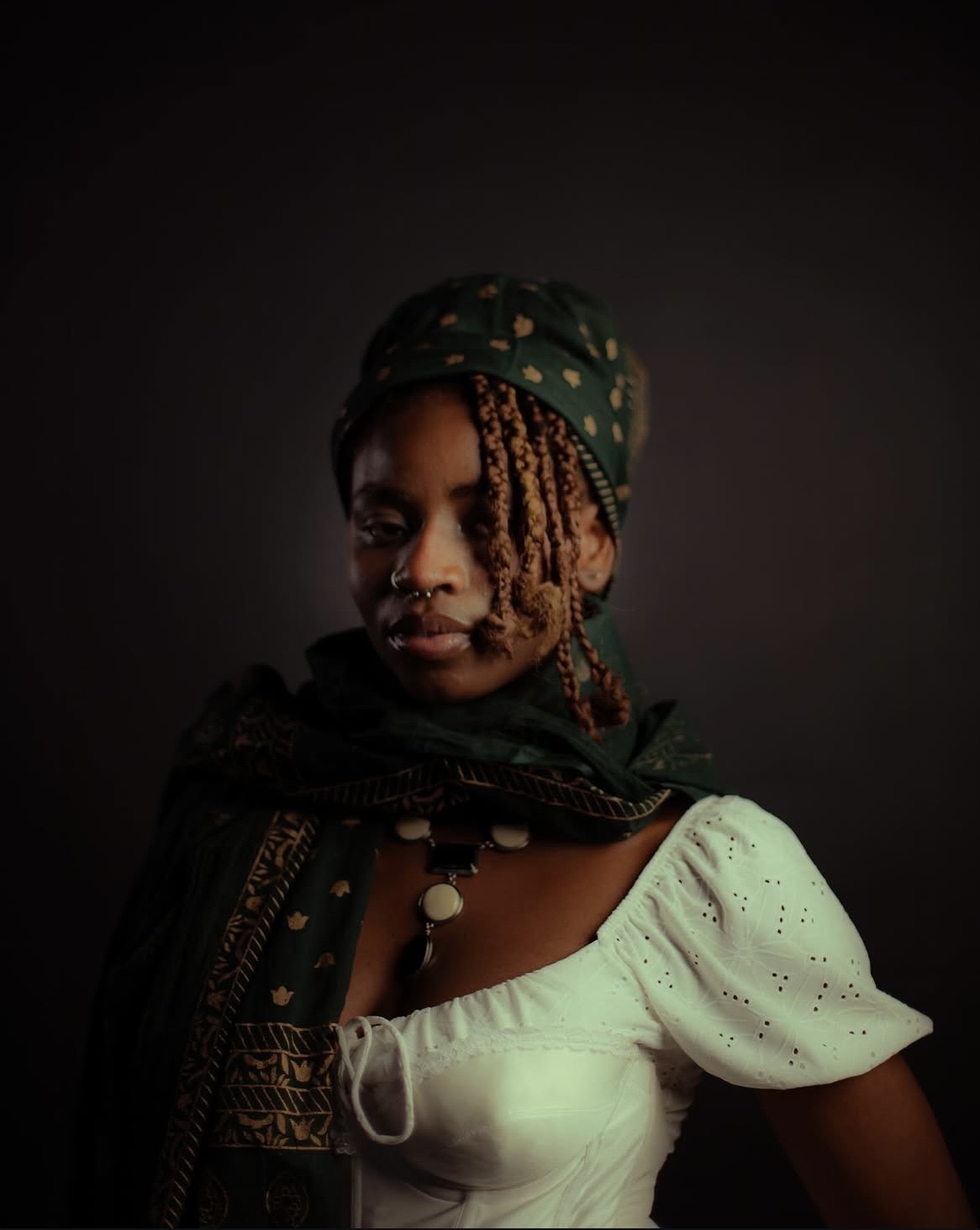 Portrait of a woman with braided hair wearing a headscarf and white dress against a dark background.