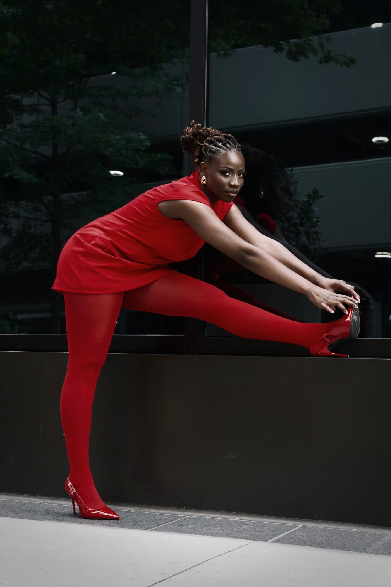 Woman in red outfit stretching her leg on a black surface outdoors, with trees and building reflections in the background.