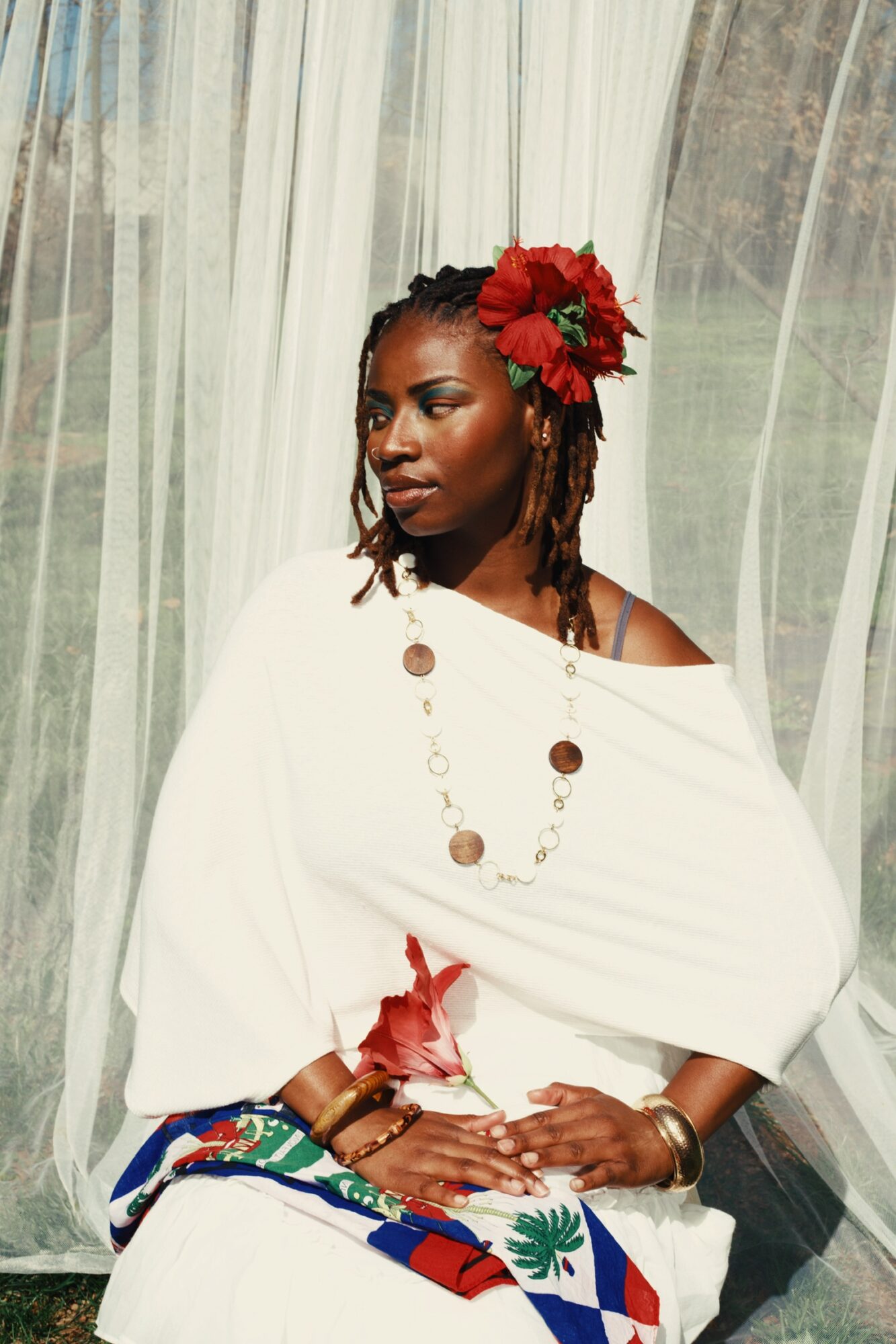 Woman with red flowers in hair, wearing white dress and jewelry, sitting in front of sheer curtains outdoors.