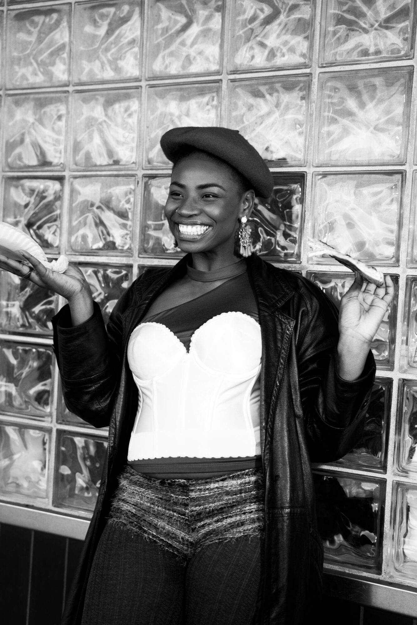 Smiling woman wearing a beret, leather jacket, and striped pants, standing in front of a glass block wall.