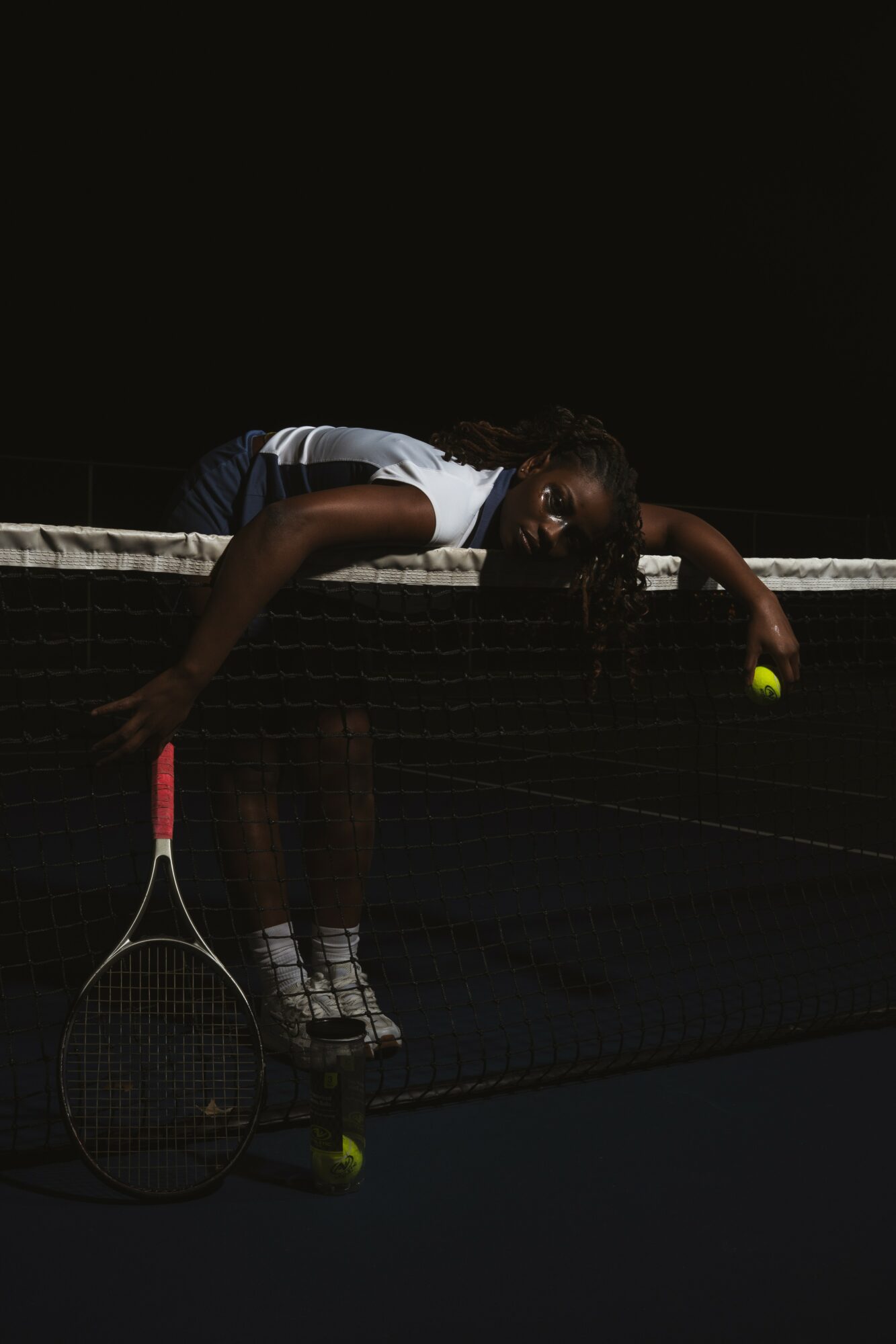 Person leaning over tennis net holding a tennis racket and a tennis ball, dark background, indoor court.