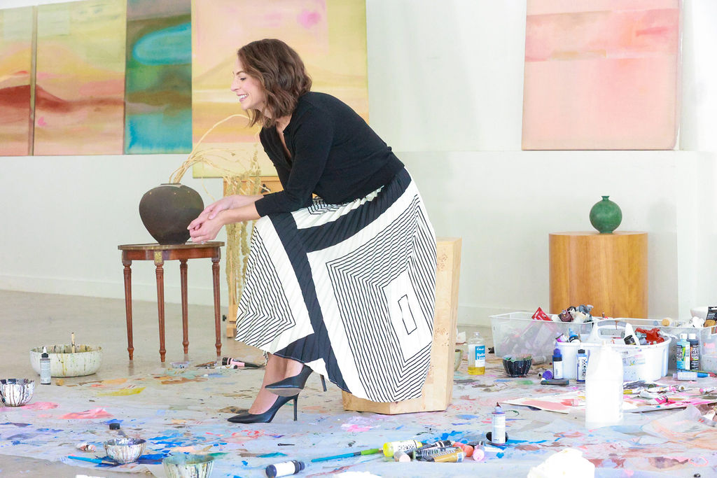 Woman in a black top and patterned skirt reaches for a bowl on a small table, surrounded by paint supplies and colorful artwork.