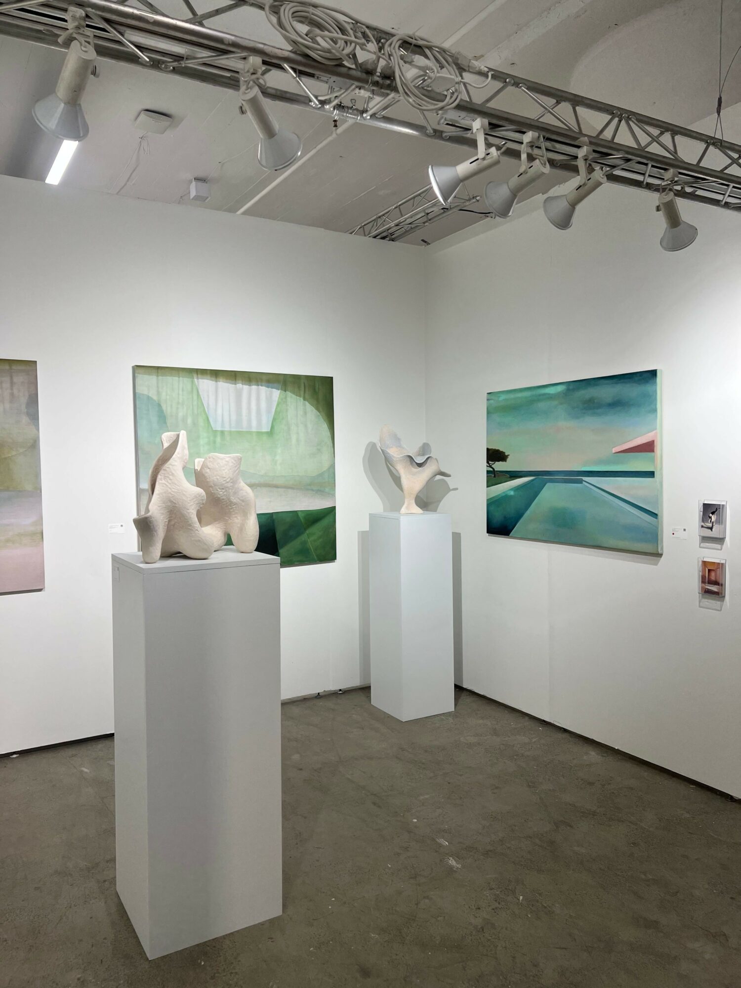 Gallery with paintings on white walls and sculptures on pedestals, concrete floor, and ceiling with lighting fixtures.