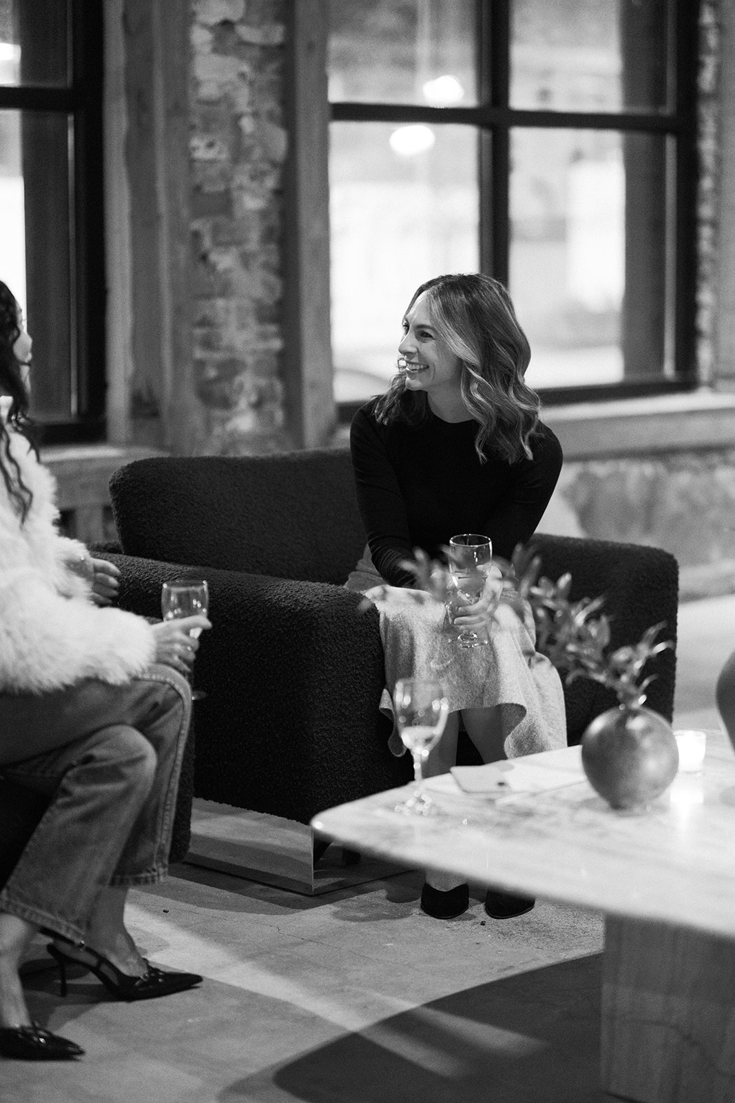 Two women sitting on a sofa and talking in a room with large windows and brick walls.