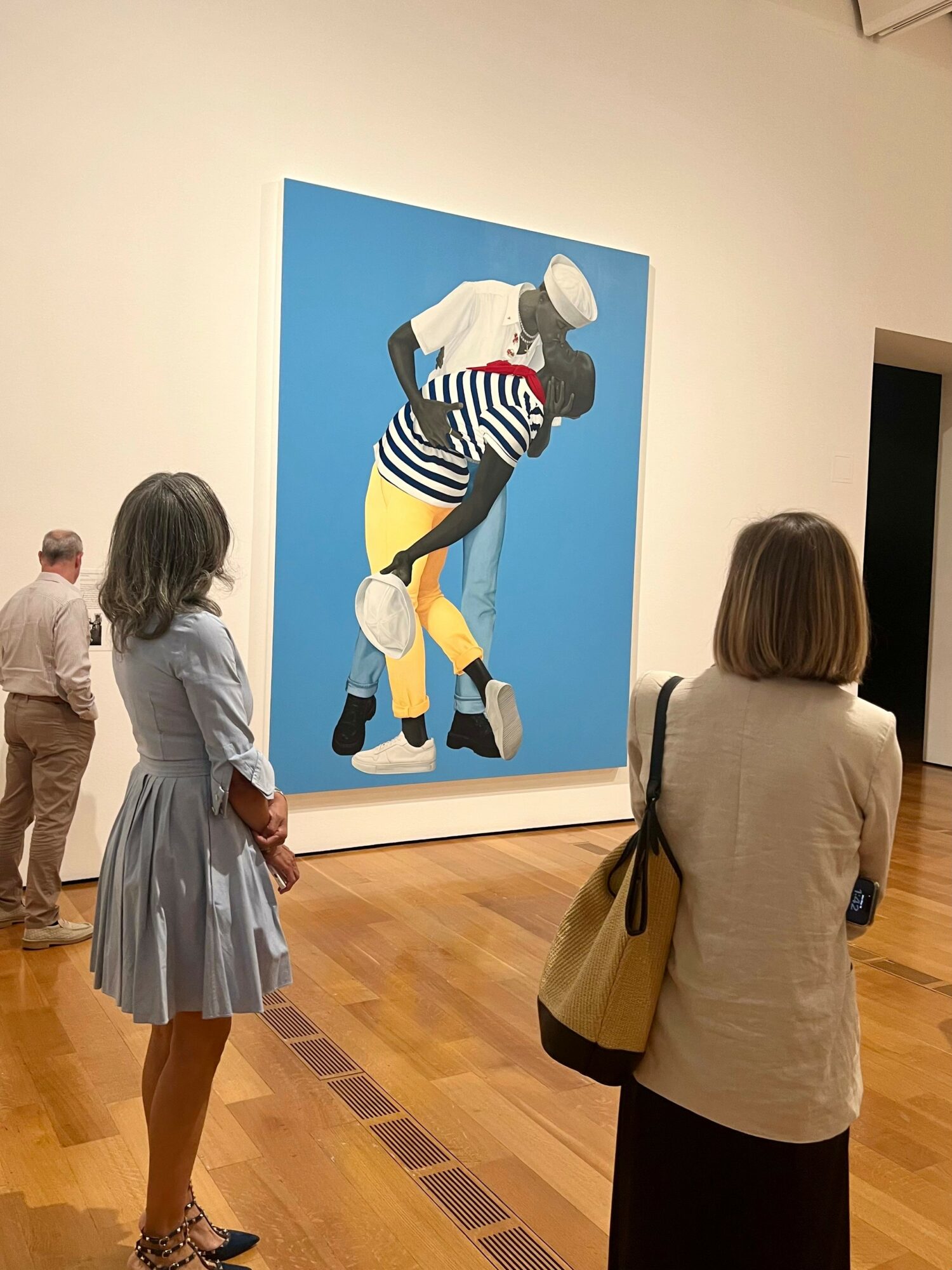 Three people observe a large colorful painting of a person with a striped shirt and yellow pants in an art gallery.