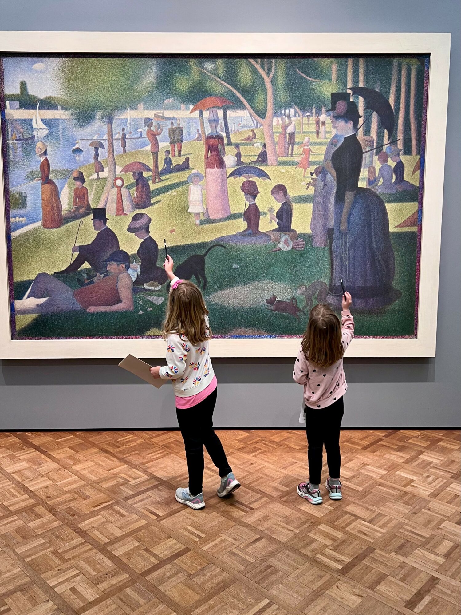 Two children stand in front of a large painting of a park scene with people, trees, and a pond.