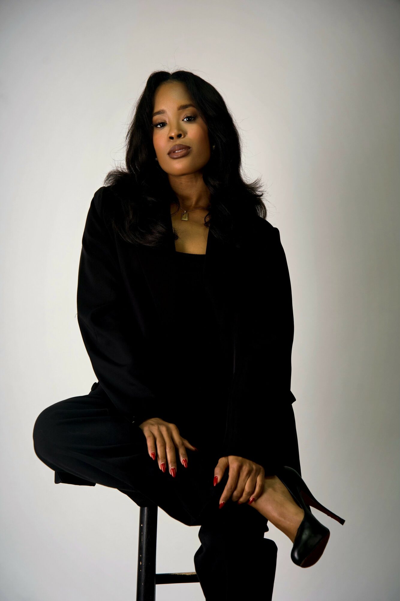 Woman with long dark hair wearing black clothing, sitting on a stool against a plain background.