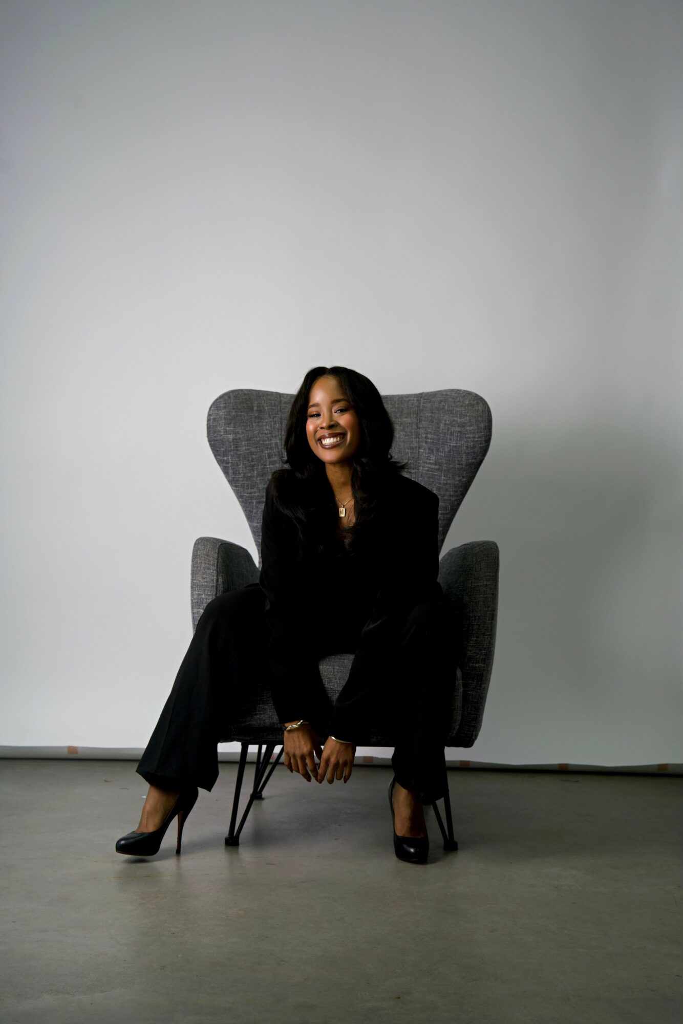 Woman sitting on a modern gray armchair, smiling, wearing black clothing, in a minimalistic room with plain white wall.