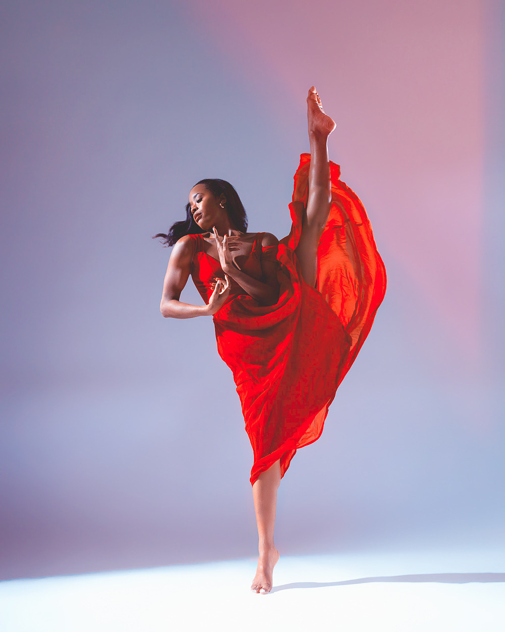 A dancer in a red dress performs a high kick with one leg extended upward, against a gradient background.