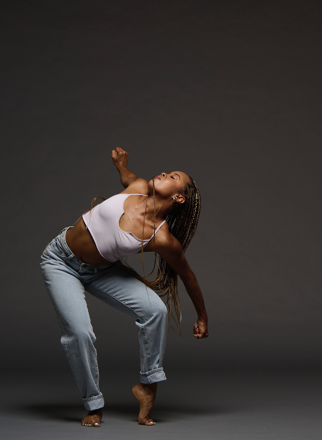 Person dancing with one arm raised and head tilted back, wearing a white top and jeans, against a dark background.