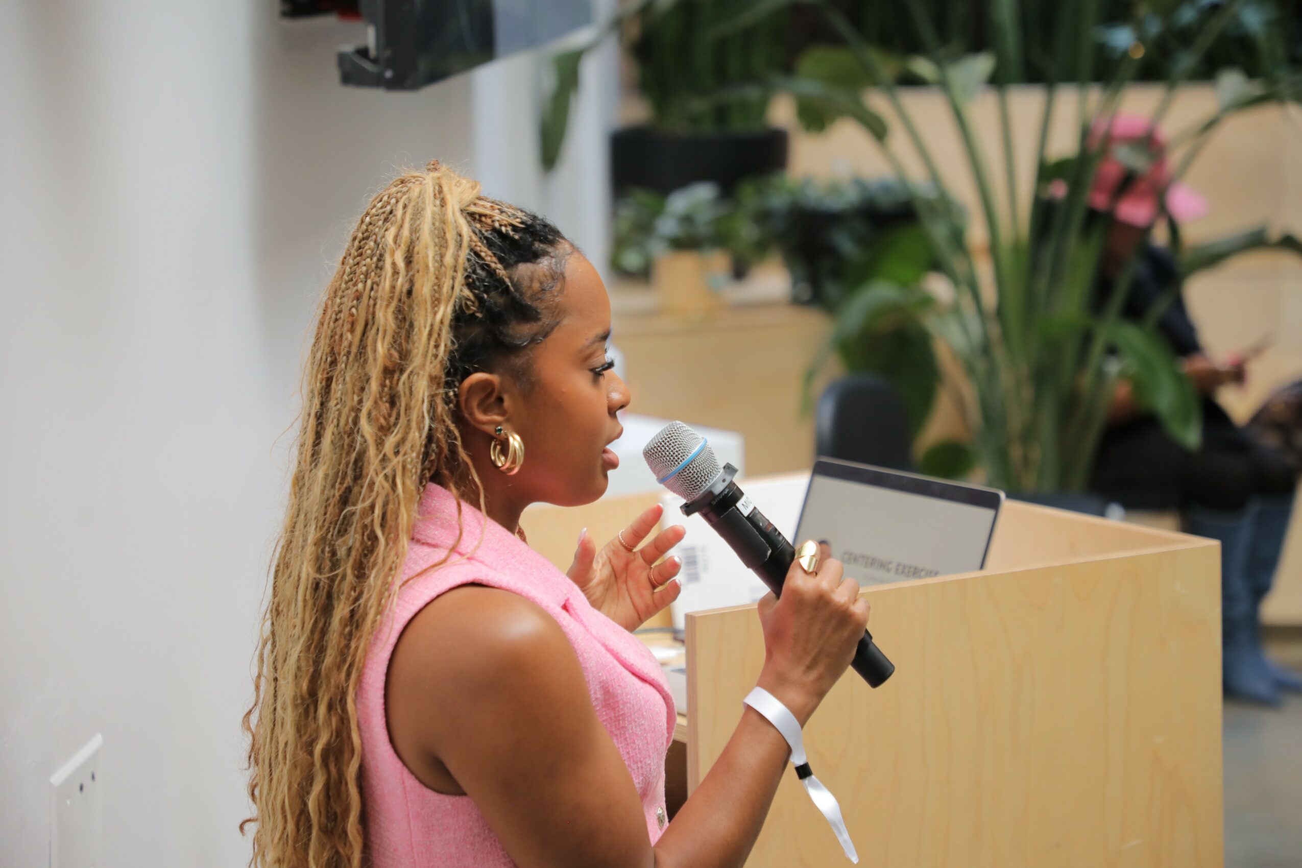 Woman with long curly hair speaking into a microphone at a podium, wearing a pink sleeveless top, in a room with plants.