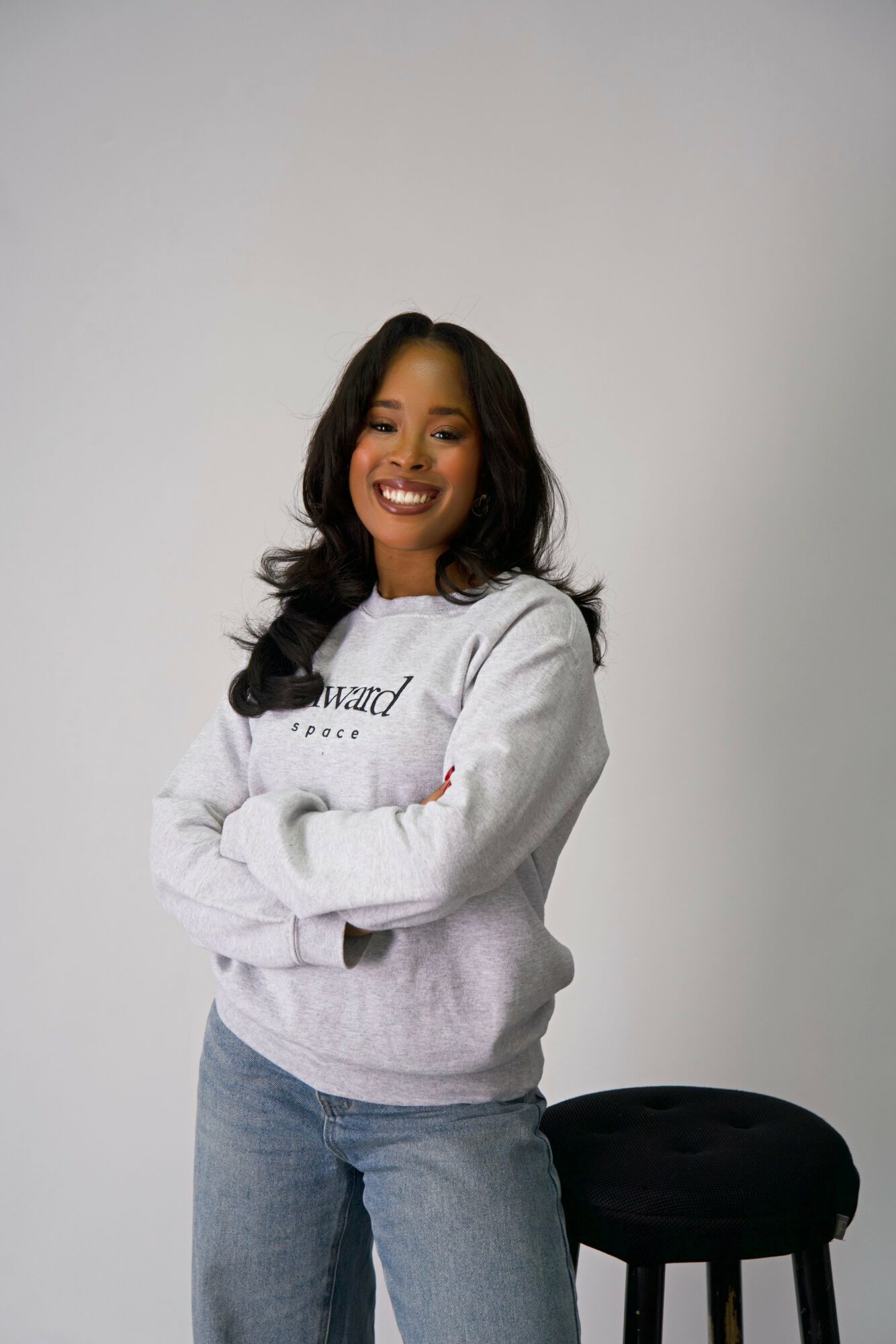 Smiling woman with long dark hair wearing a light gray sweatshirt and jeans, standing with arms crossed next to a black stool.