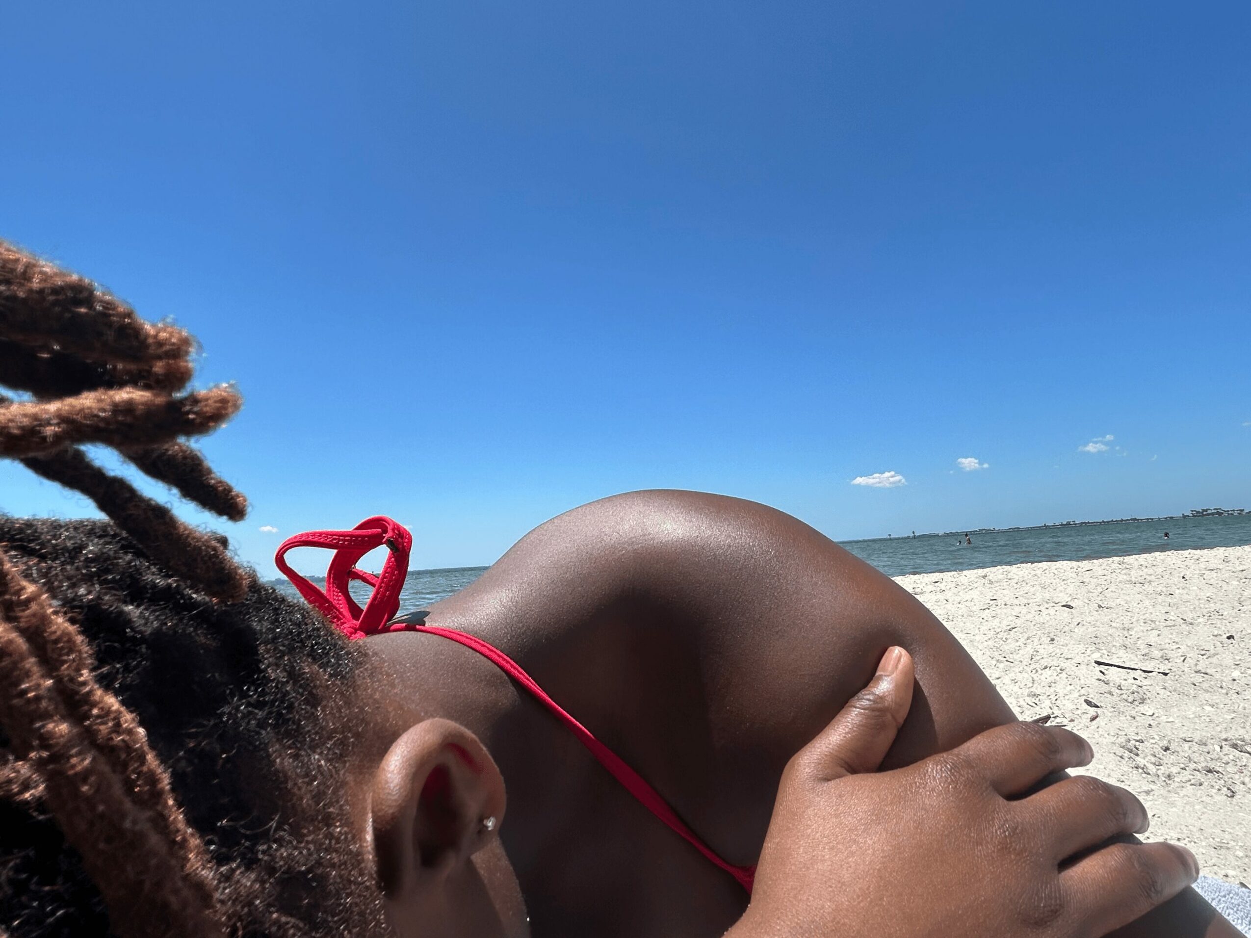 Person lying on a sandy beach with a clear blue sky, wearing a red swimsuit, hand resting on their back.