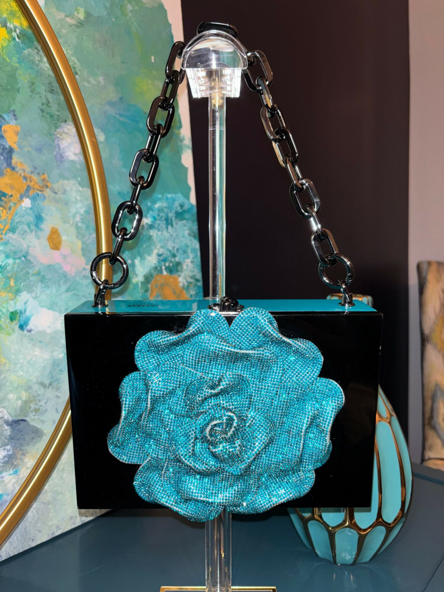Decorative object with a blue, textured rose on a black rectangular base, chain and rod support, against a colorful background.