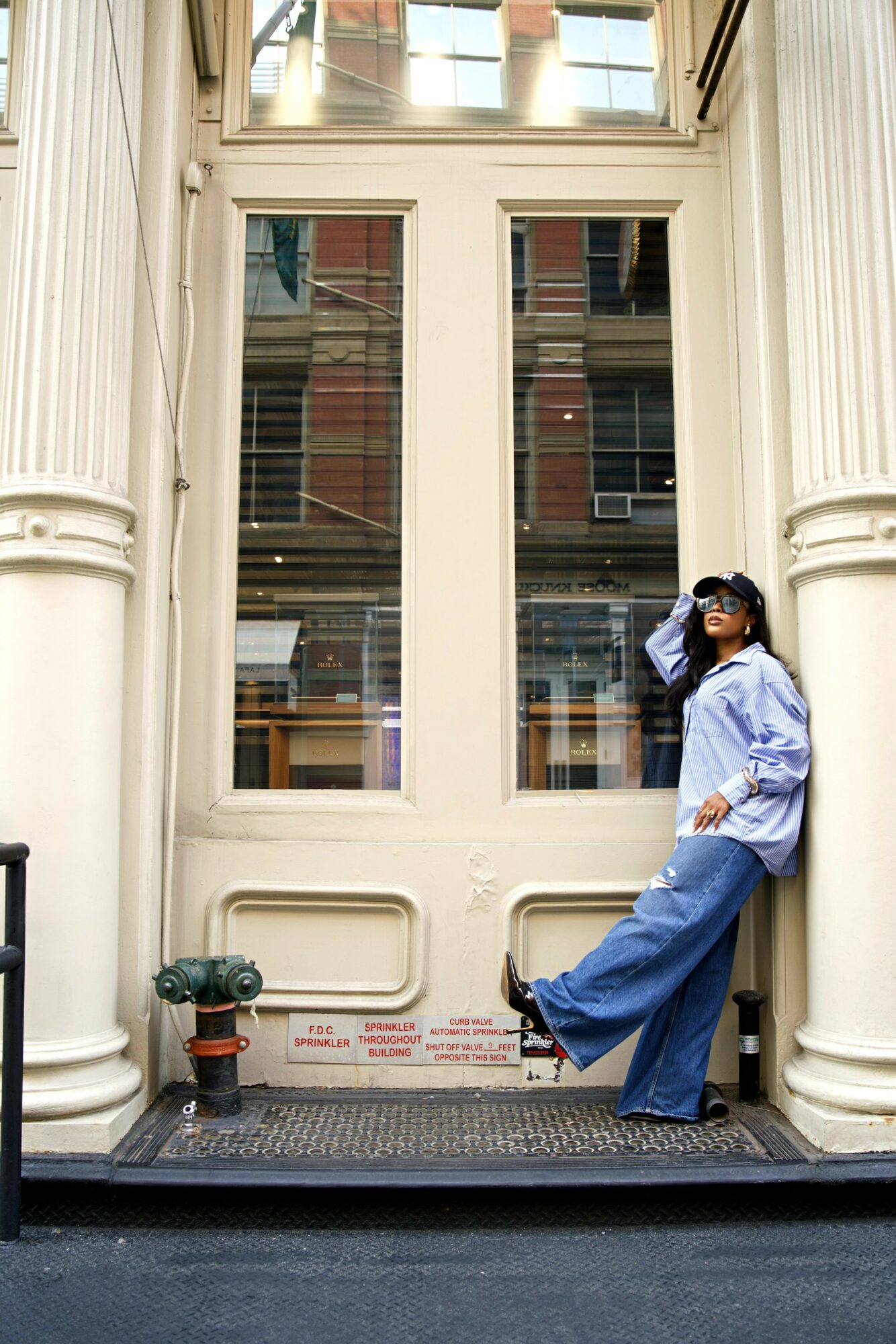 Person with sunglasses and long hair, wearing a light blue shirt and wide pants, leaning against a large door with columns.