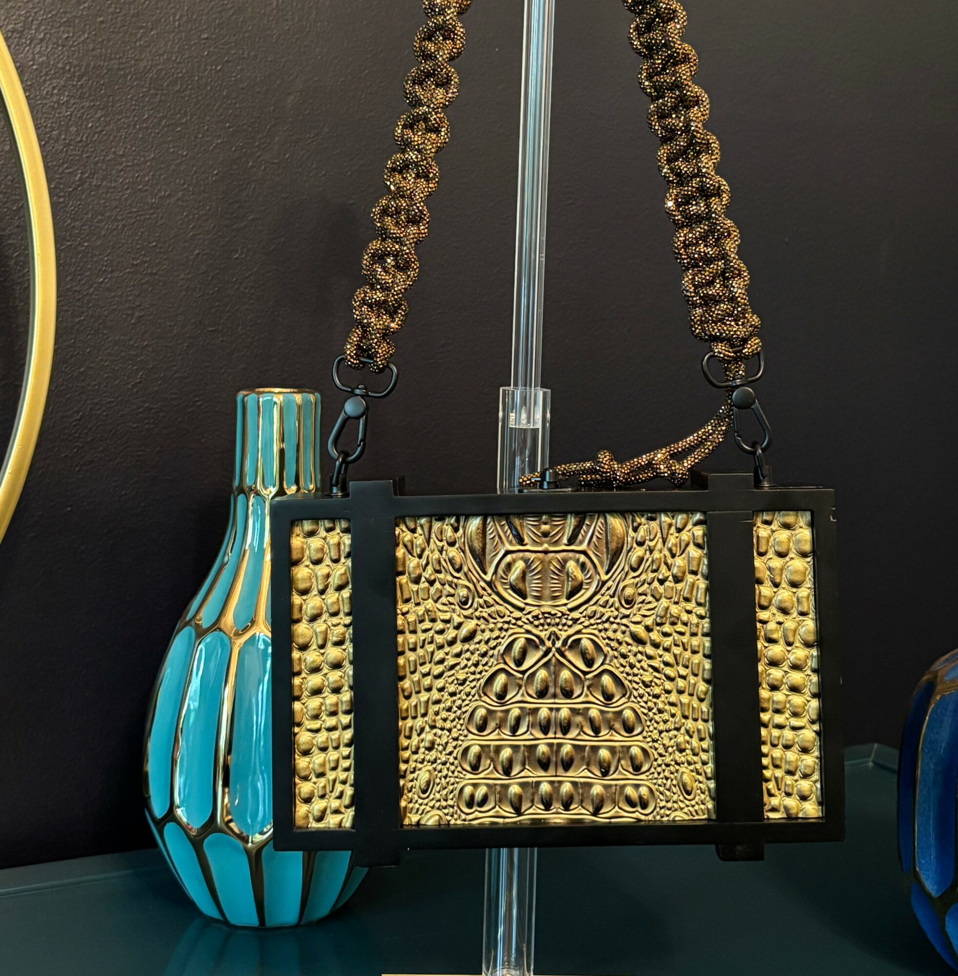 Black and gold handbag with a chain strap, displayed on a stand, with a blue and black vase nearby.