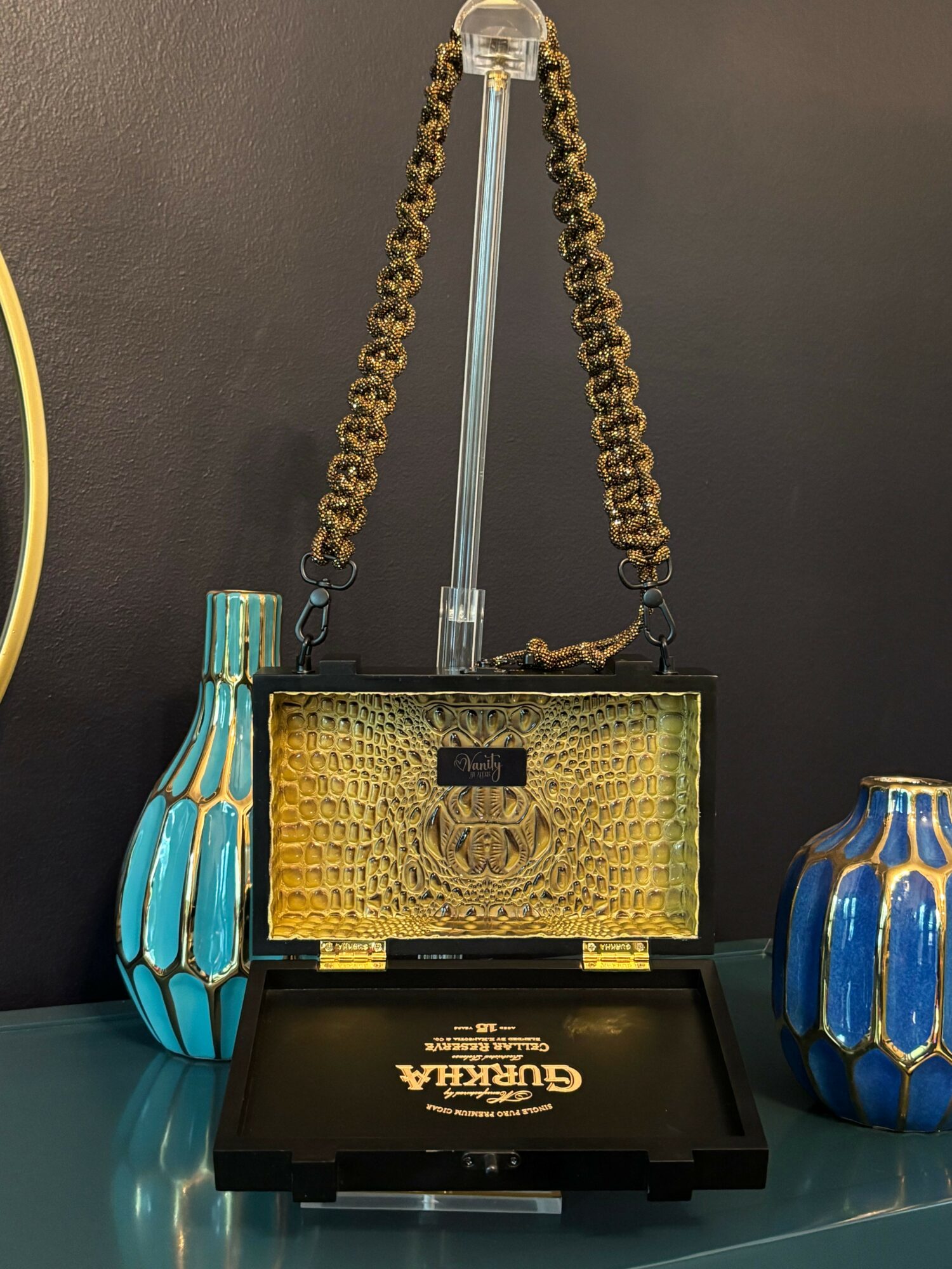 A gold-colored handbag with a chain strap, displayed on a black surface with blue and gold decorative objects nearby.